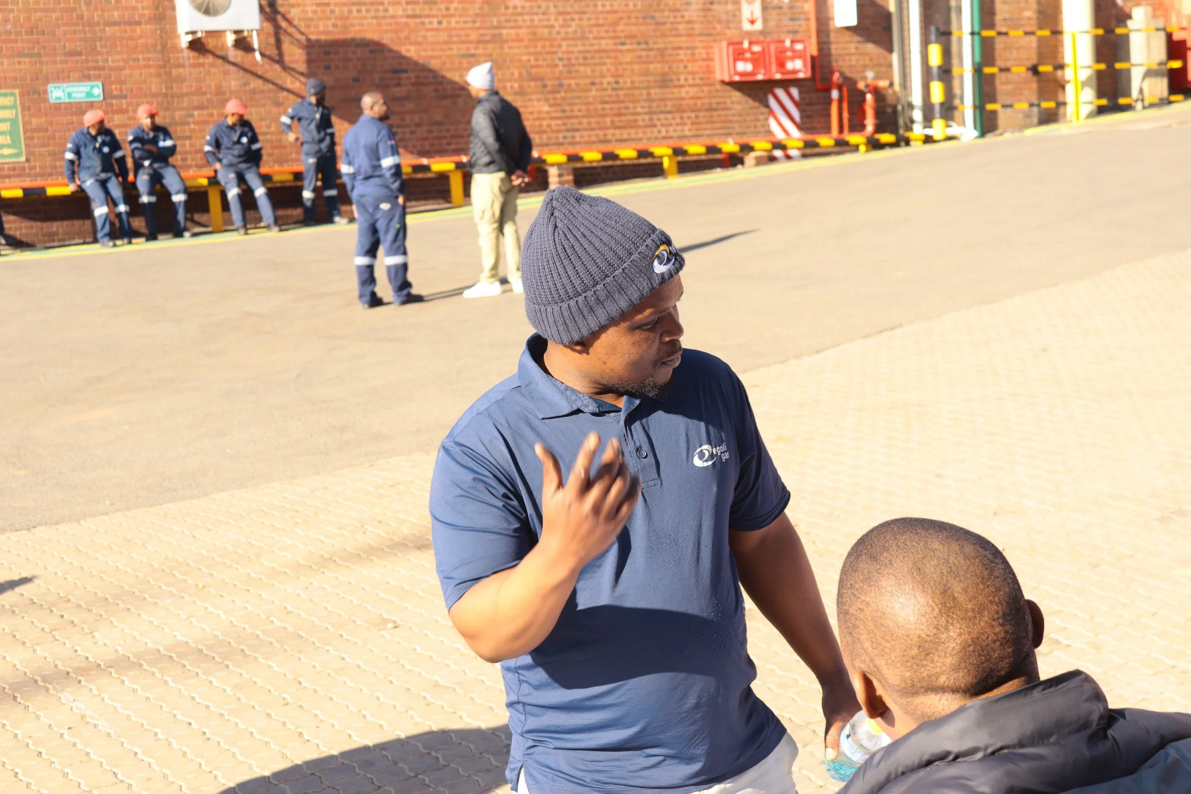 A man wearing a gray beanie and blue polo shirt appears to be talking to a child in a black jacket and holding a water bottle, outdoors near a brick building with a group of people sitting on a yellow and black barrier in the background.