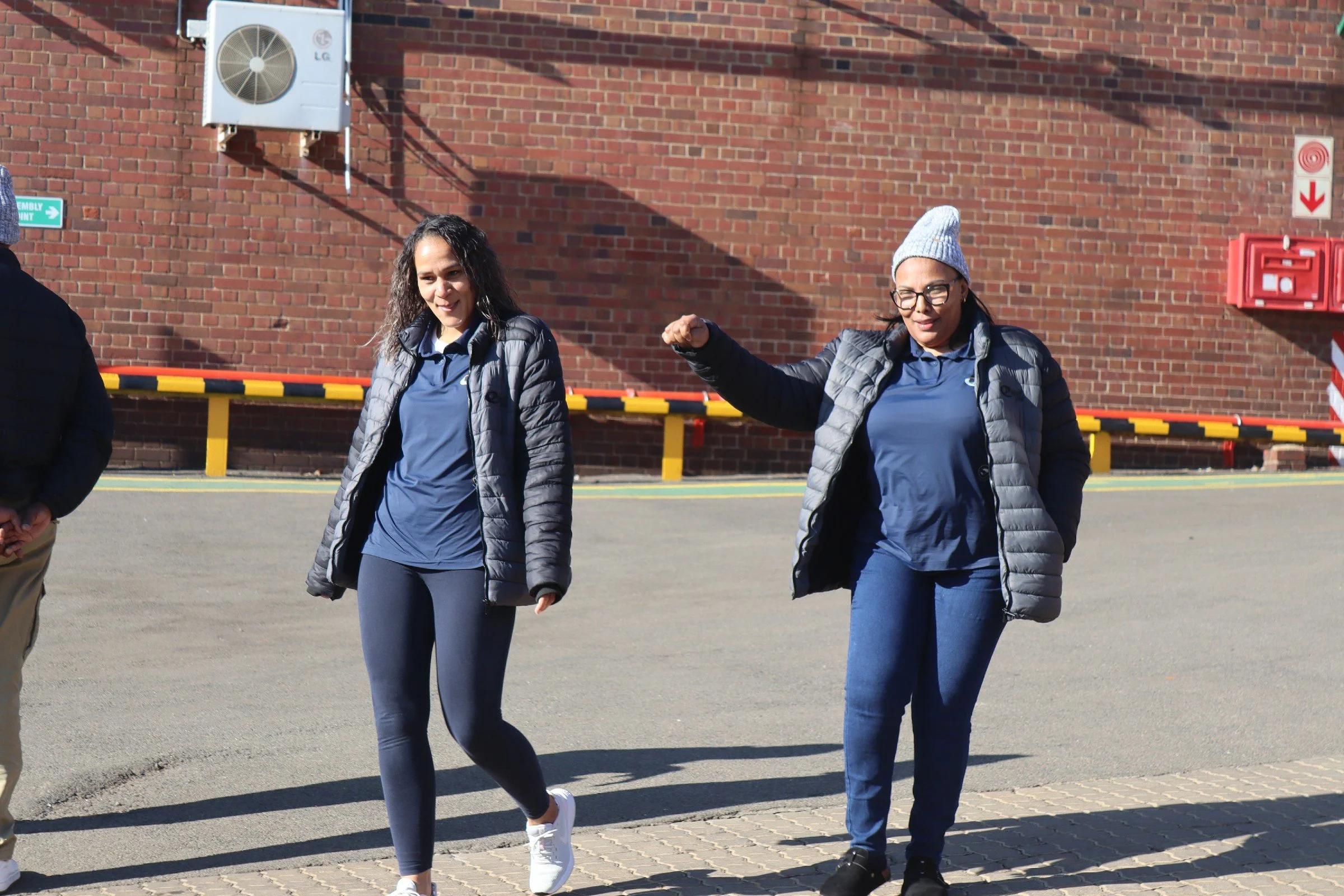 Two women walking outdoors in front of a brick wall, both wearing blue shirts, dark jackets, and beanies, with one woman making a gesture with her hand.