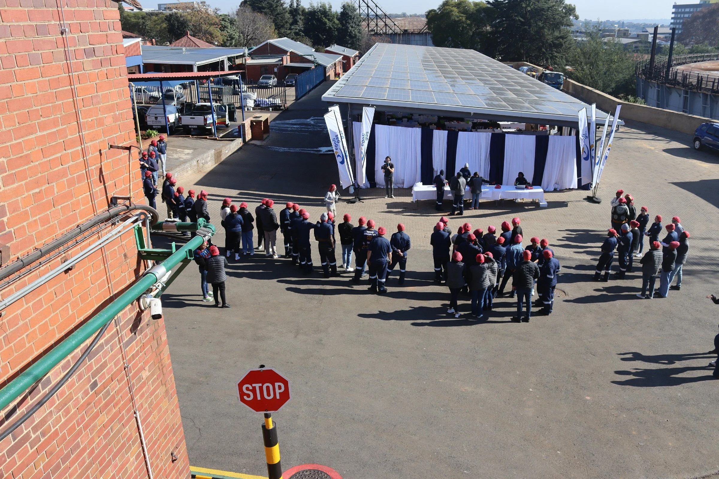 A large group of people wearing red hats standing in line outdoors near a white tent with blue and white banners, with a brick building on the left and trees in the background.