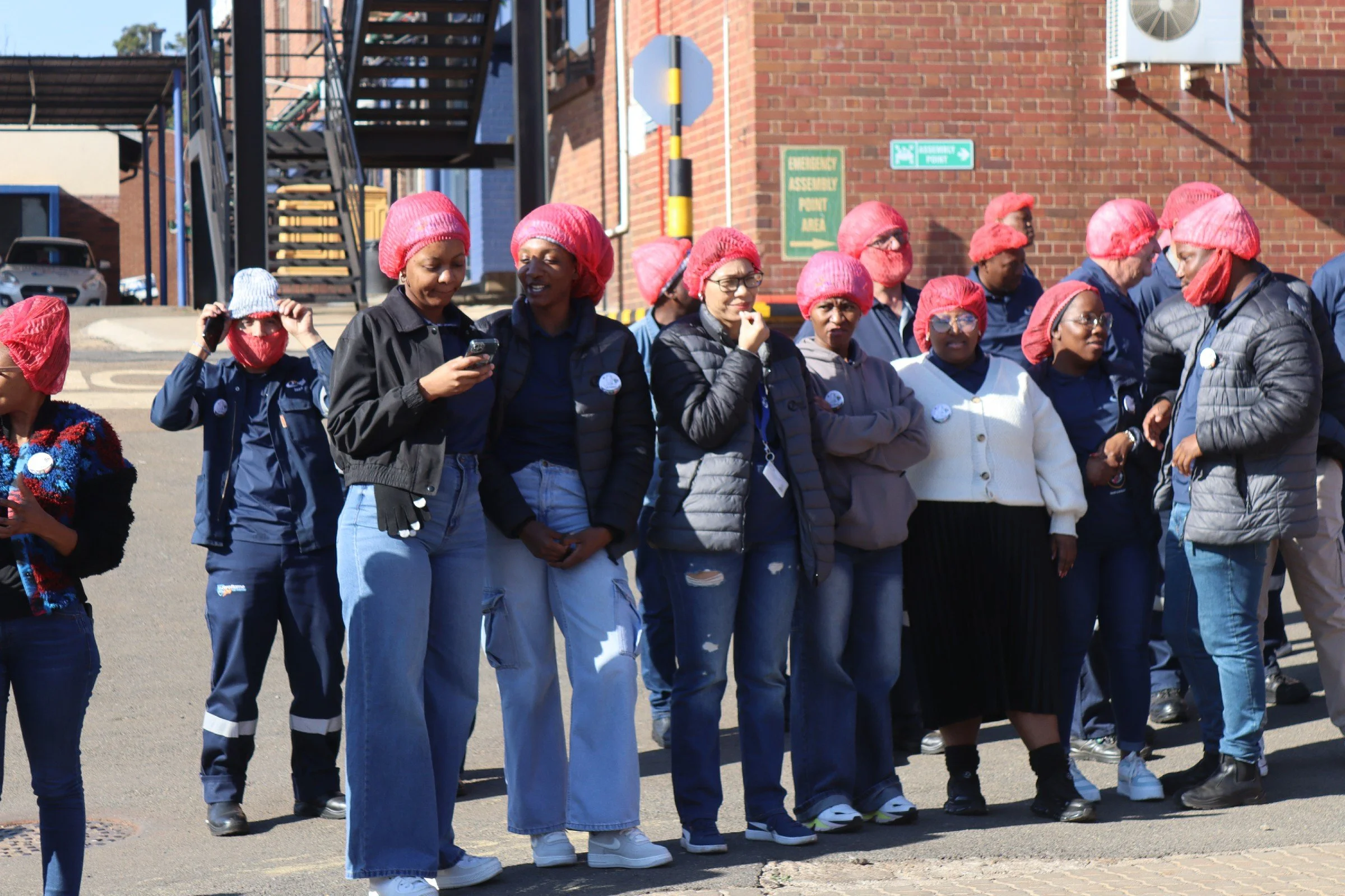 A group of people wearing red hair covers or caps standing outdoors. Most are dressed in dark or casual clothing, some are wearing reflective patches or badges. The scene takes place near a brick building with parking signs, a staircase, and parked c
