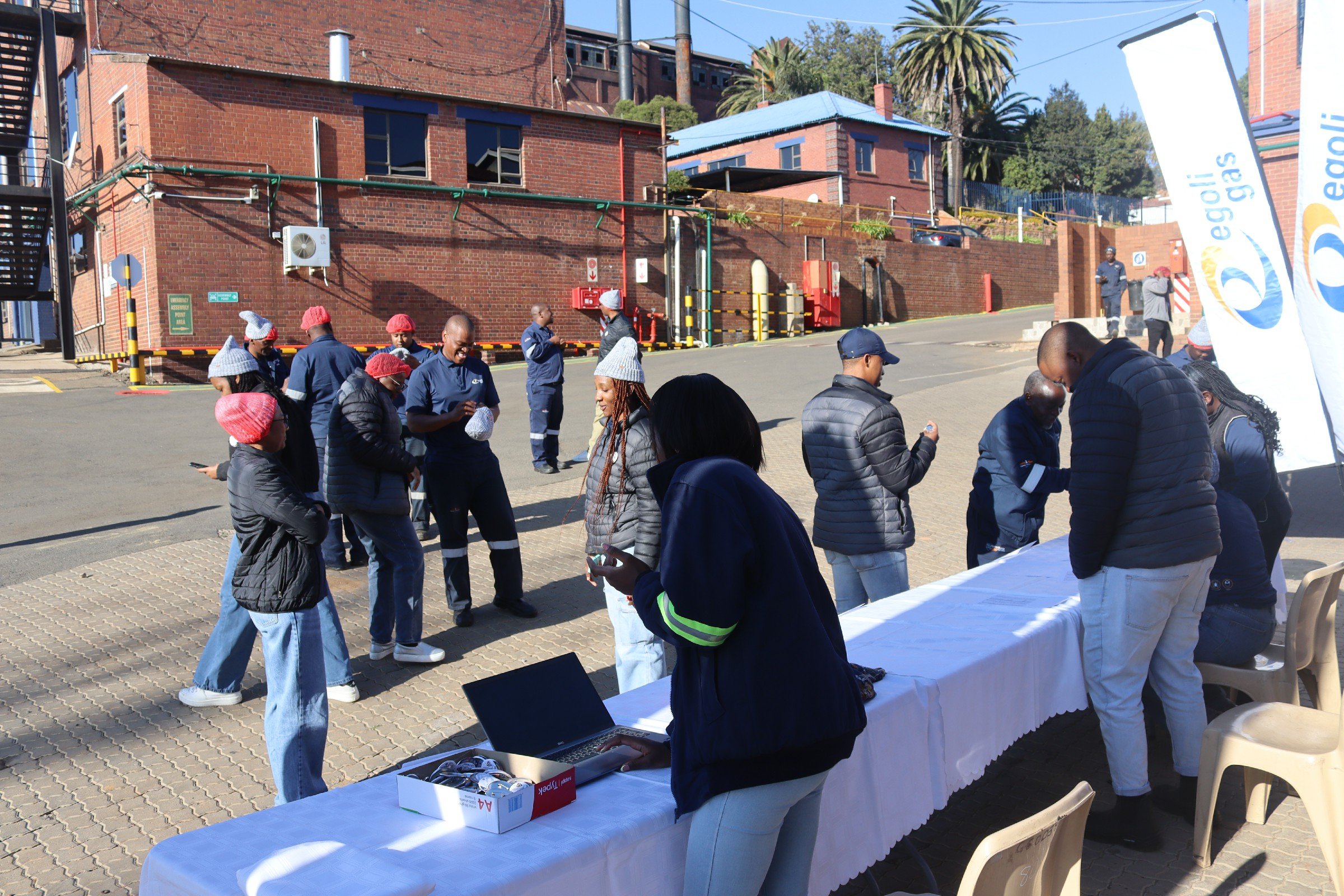 People gathered outdoors in front of a brick building with trees in the background, near a table with a white tablecloth and electronic devices, some wearing hats and jackets, against sunny weather.