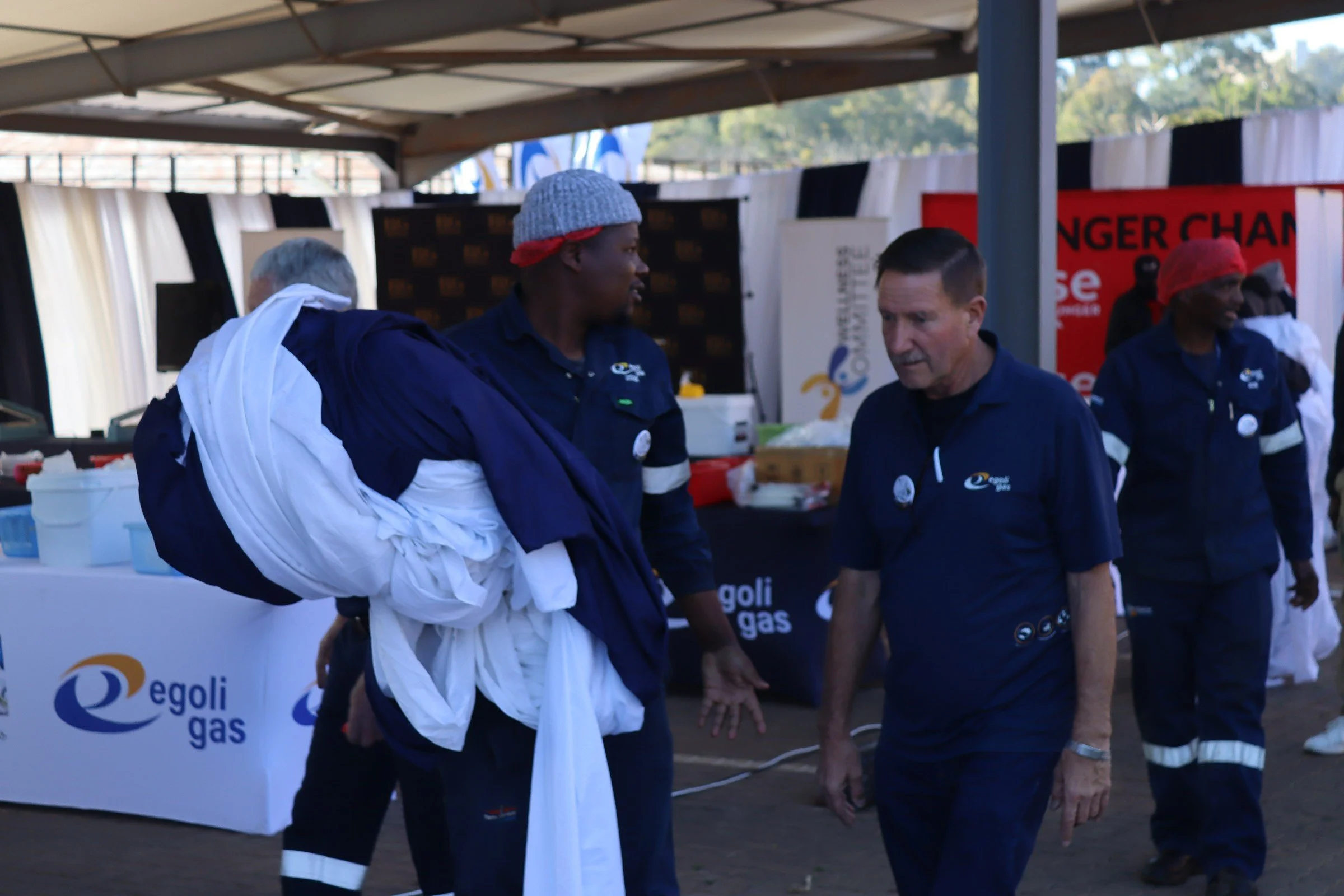 Two men in navy blue shirts are talking to each other at an outdoor event, with a group of people in similar attire behind them. There are banners and a table with supplies under a canopy.