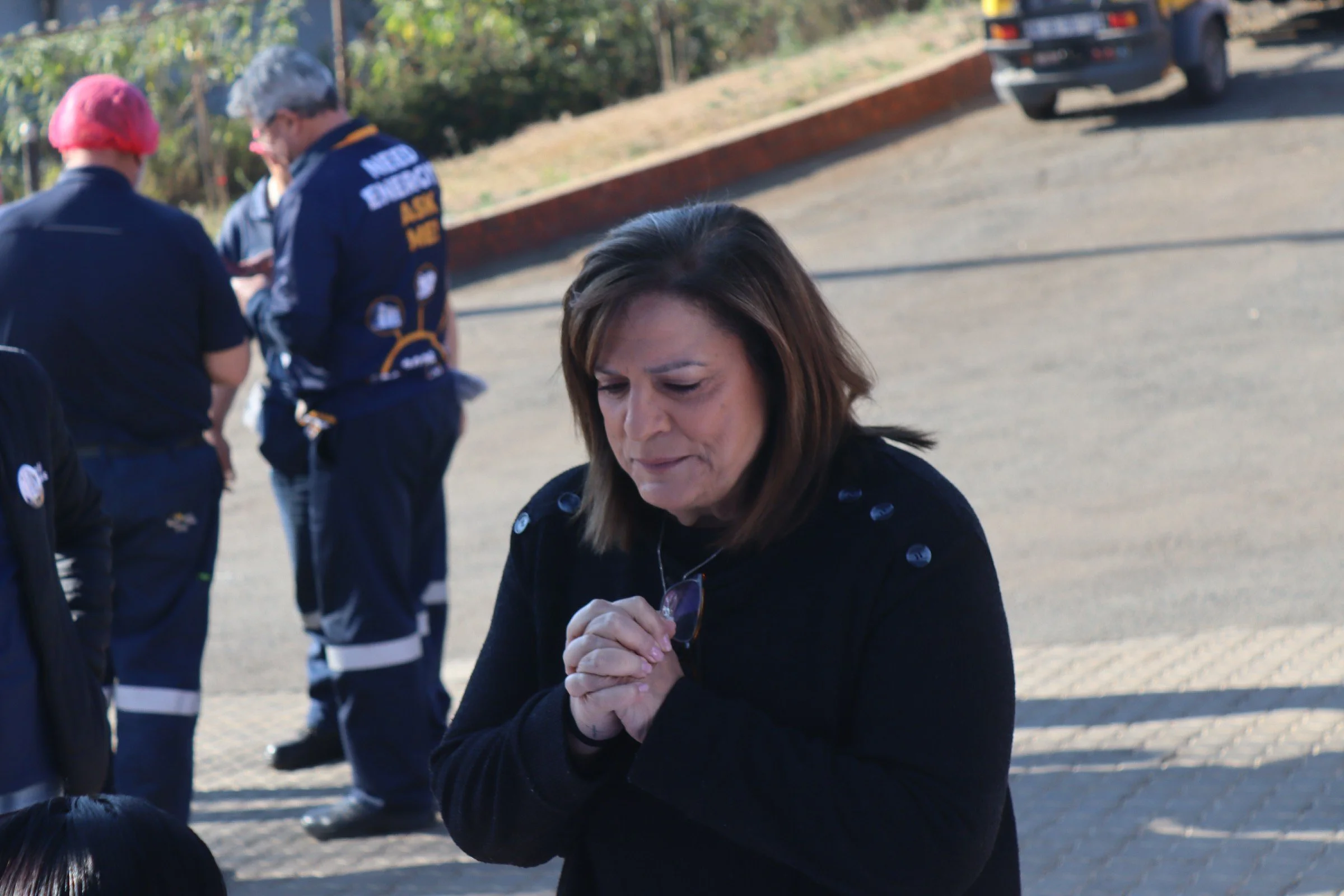 A woman with brown hair in a prayer position with her hands clasped, eyes closed, and a solemn expression. She is wearing a black jacket. In the background, there are emergency responders in navy uniforms with reflective stripes and helmets, standing