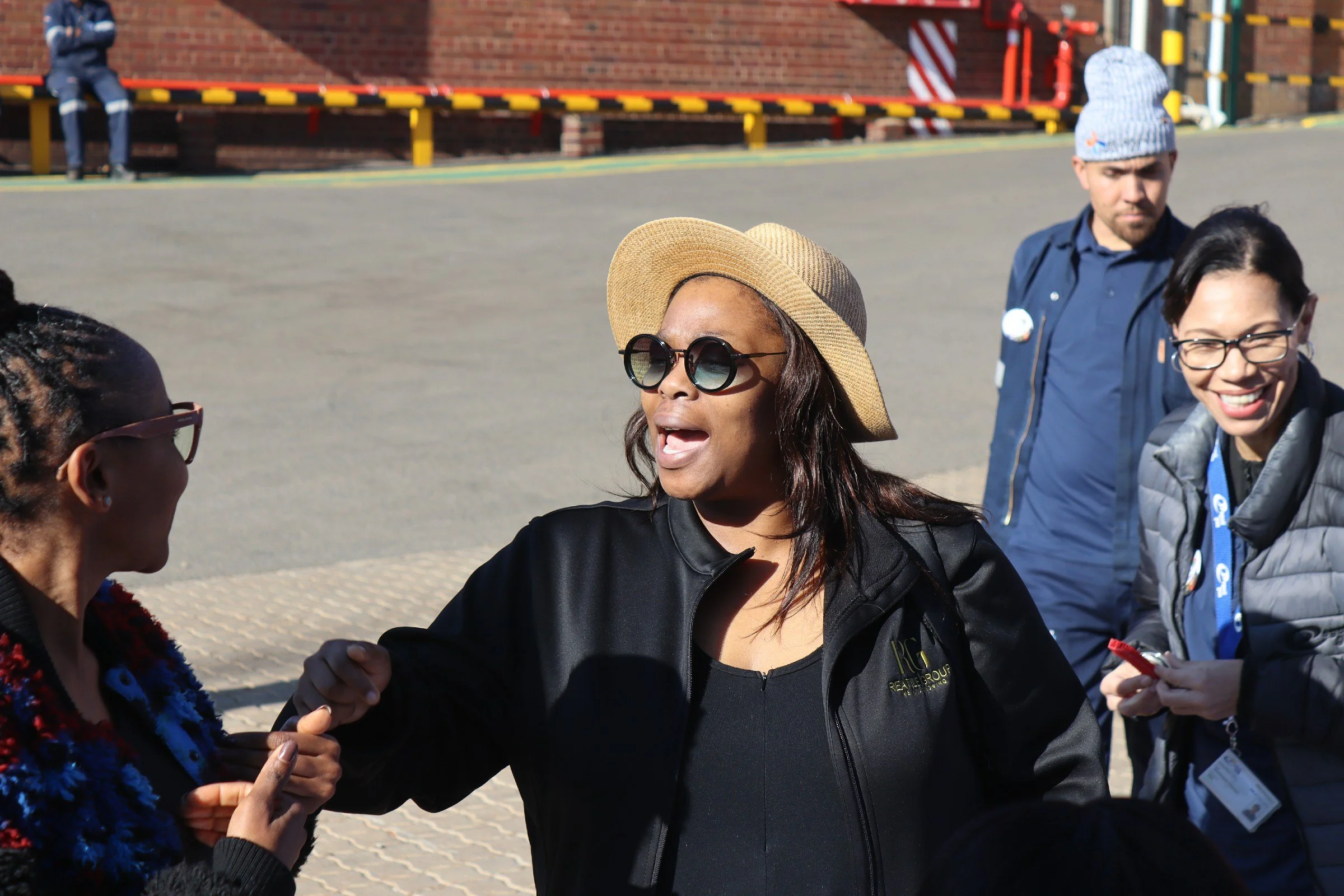 A group of diverse people engaging in a conversation outdoors, with some smiling and gesturing. A woman in the center wears sunglasses and a sunhat, and is speaking to a woman on the left, while others in the background observe and participate.