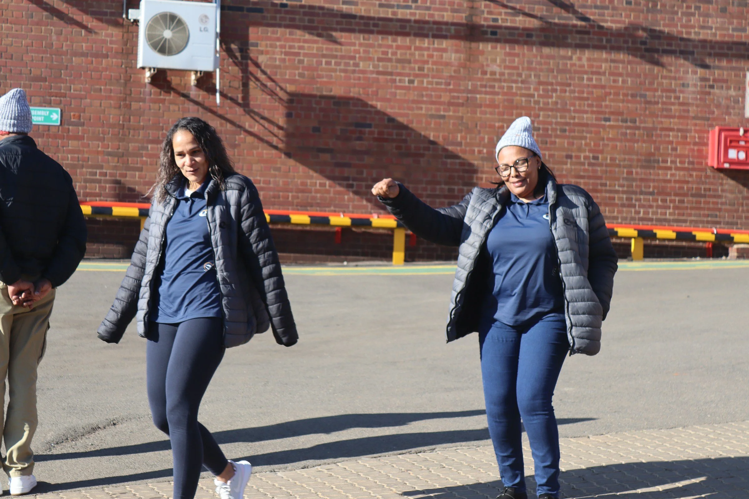 Group of four women walking outdoors in front of a brick wall, two women are visible smiling and wearing gray beanies and gray jackets, the women are dressed in casual athletic clothing.
