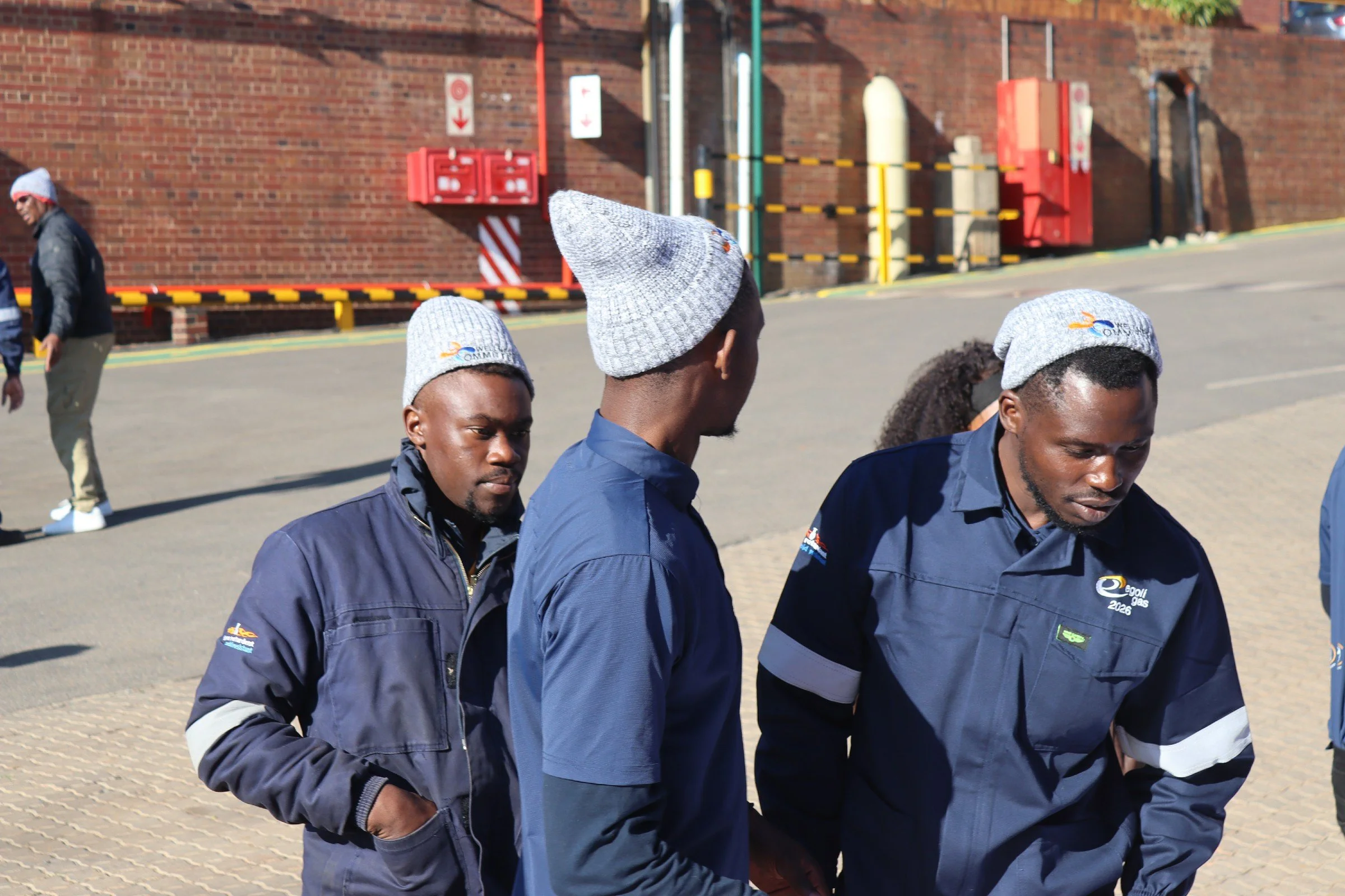 Group of workers walking outdoors near a brick wall with safety signs and equipment, wearing jackets and beanies.
