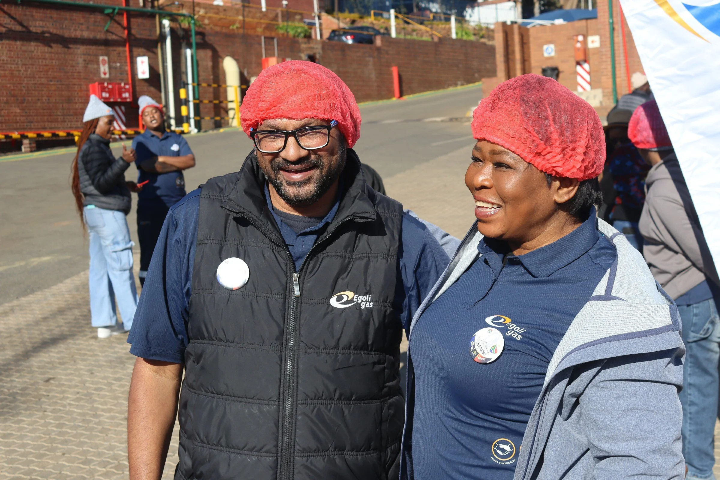 Two people smiling and wearing red hair nets, standing outdoors with a brick wall and other people in the background. They are wearing navy blue jackets with logos and buttons.