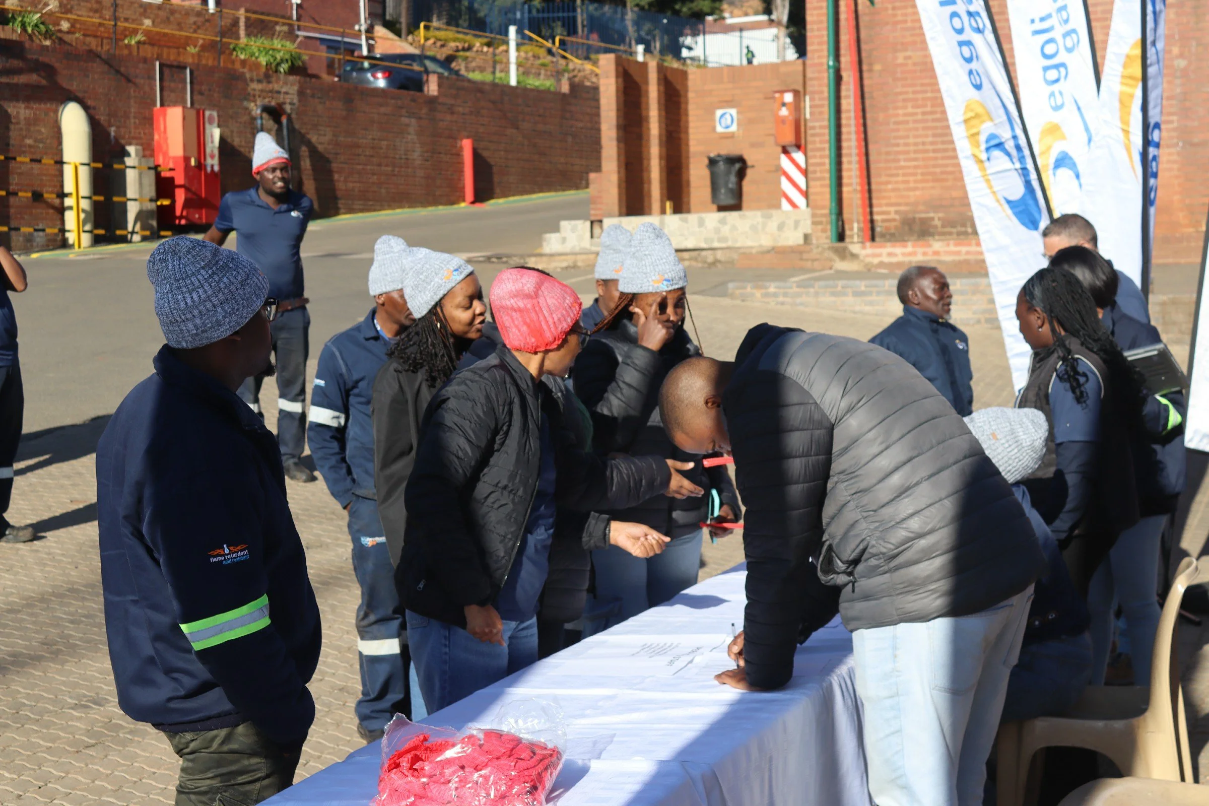 Group of people gathered around a table outdoors, some wearing matching blue uniforms with gray knit hats, under a sunny sky.