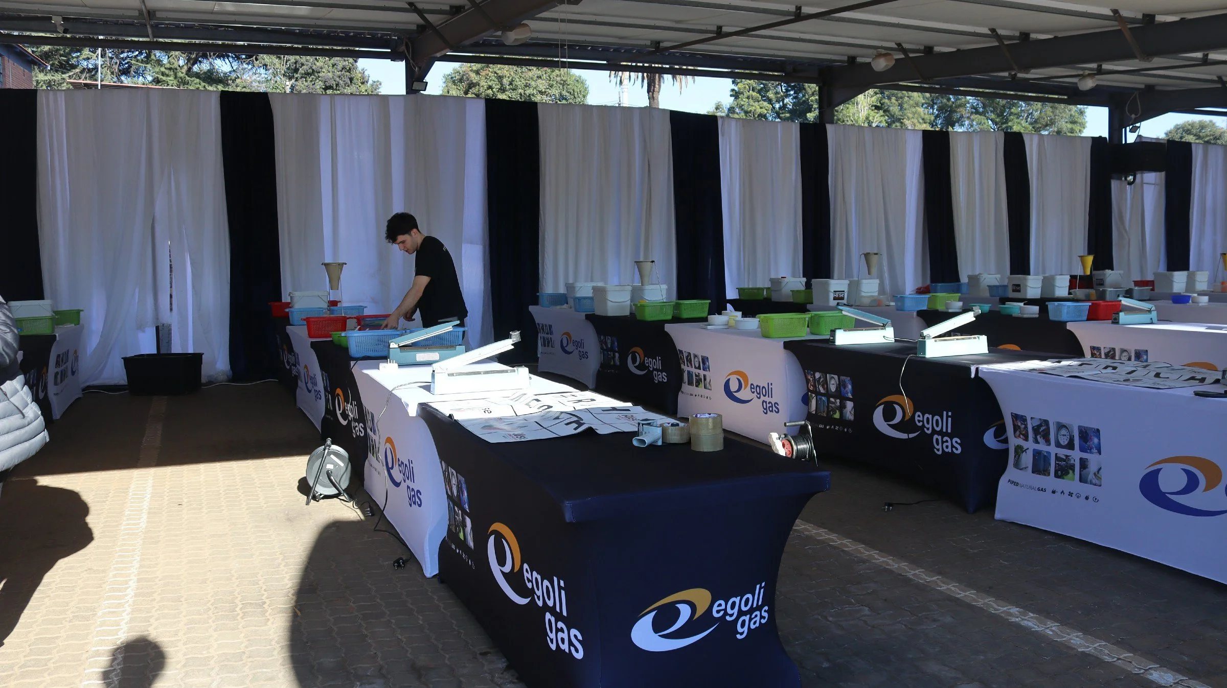 A man setting up tables at an outdoor event under a metal roof, with tables covered in black tablecloths and white banners displaying the Egoli Gas logo, with various items on the tables.