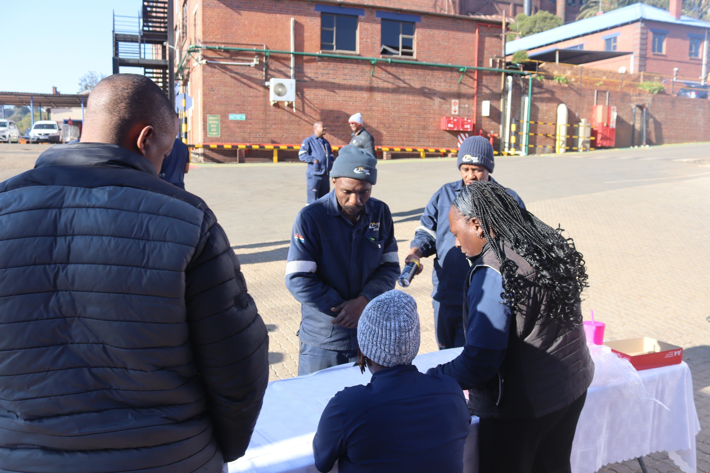 Group of people gathered around a table outdoors, engaged in a discussion or activity, with a brick building and parked cars in the background.