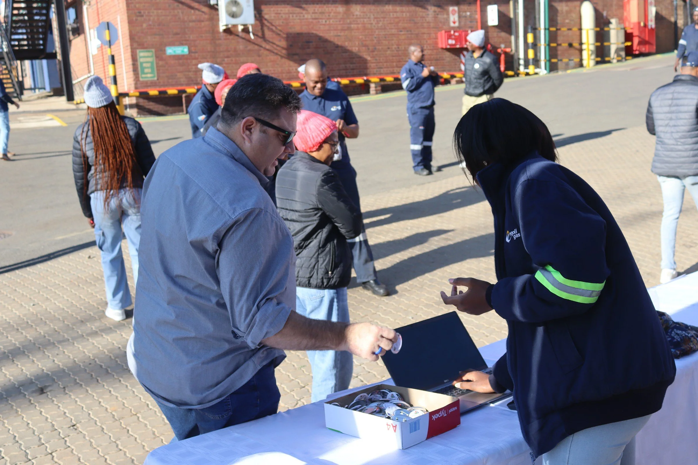 People gather around a table with a shoebox filled with medals or buttons outside a brick building, with some wearing warm clothing and hats.