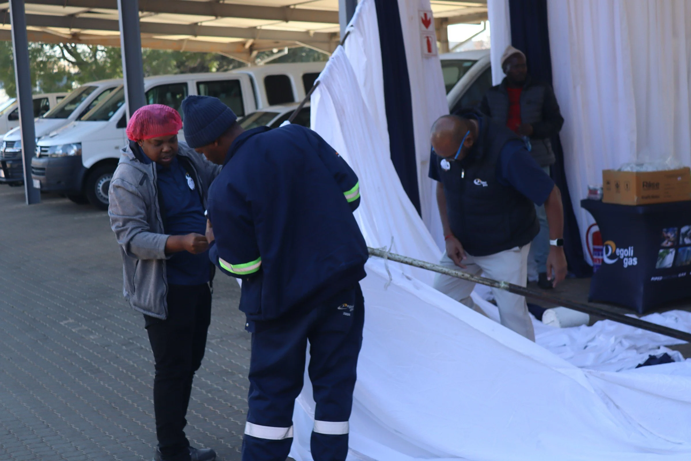 Four people are gathered around a white tent outdoors, some examining a paper or document. Three men are wearing dark jackets, with one wearing glasses and a face mask. A woman is wearing a bright pink head covering and a gray jacket. Several white v