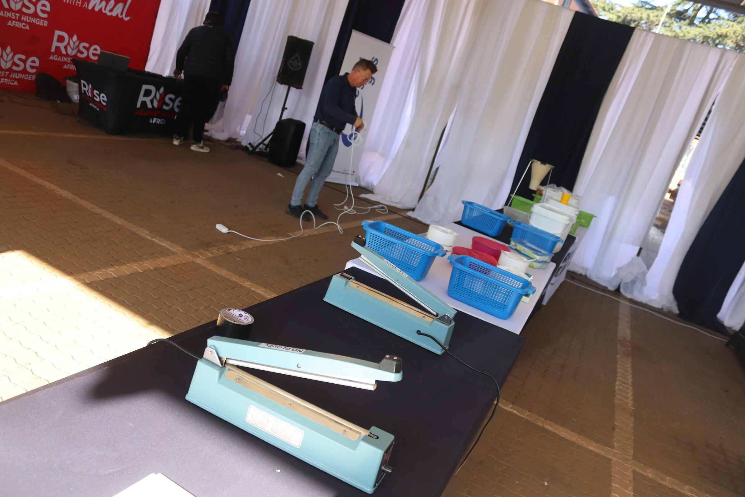 Tables with blue plastic binders, a paper cutter, and other office supplies in an indoor event space with white and black curtains.