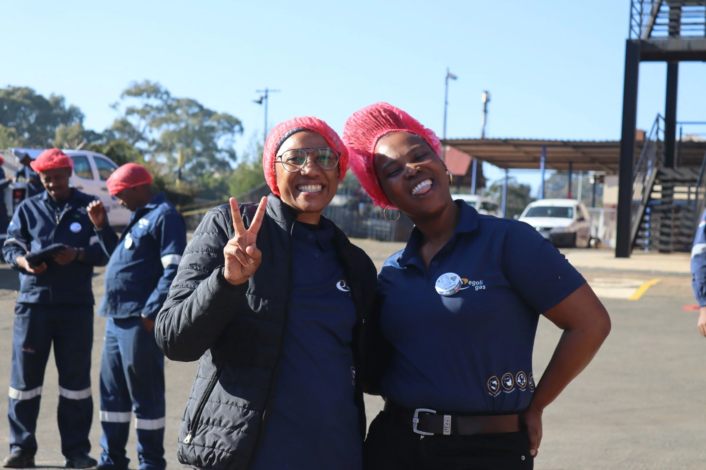 Two women smiling and making a peace sign pose for a photo at an outdoor event, wearing matching pink hairnets and navy uniforms with logos, while several others in the background also wear pink hairnets and uniforms.