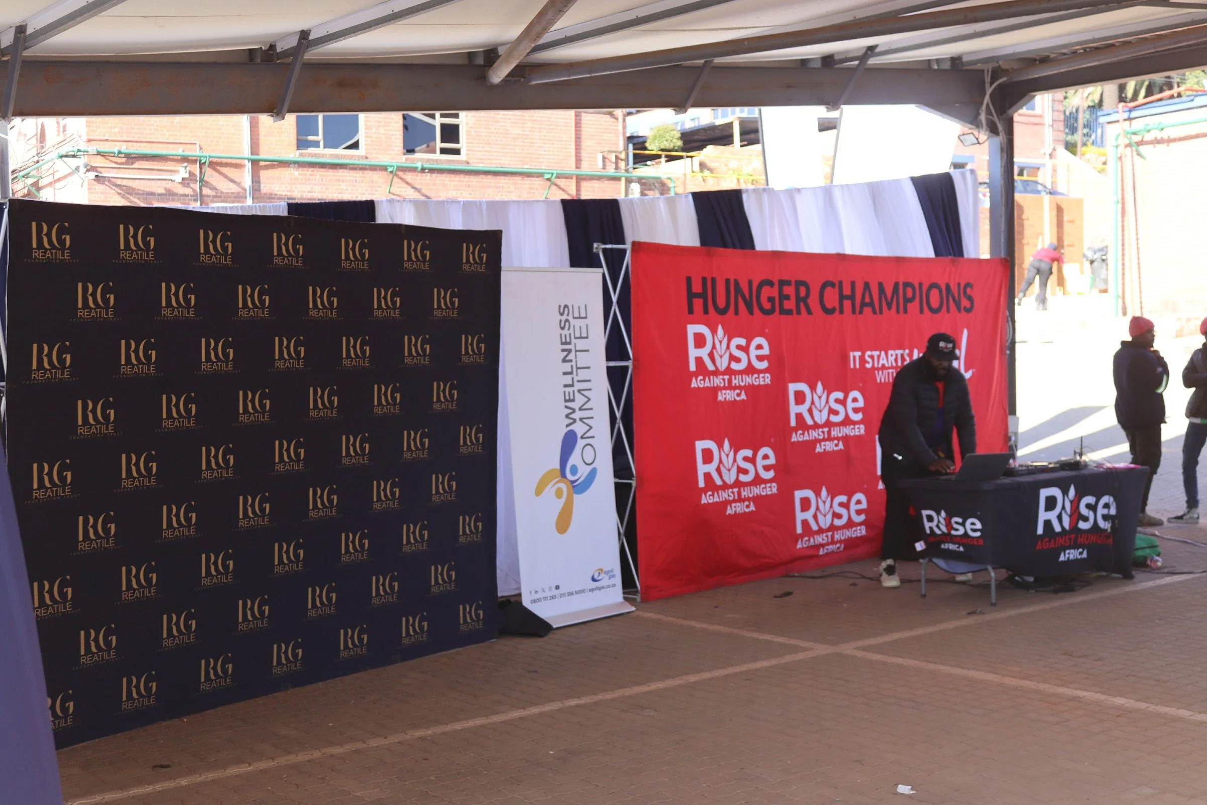 A stage setup for an event with banners, including a black banner with gold 'RG Reatile' logos and a red banner that reads 'Hunger Champions Rise Against Hunger Africa.' There are people in the background, some wearing hats, and a person at a DJ boot