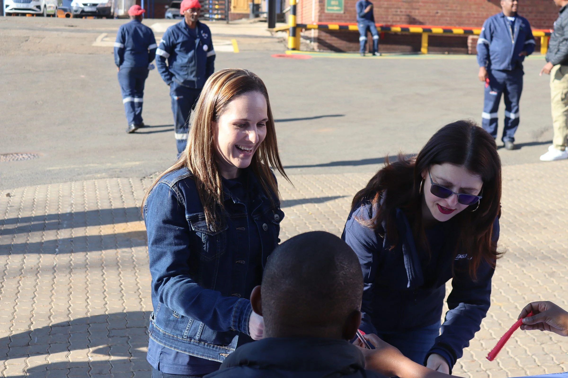 Women engaging in an outdoor activity, smiling and interacting with children. Several people are standing and walking on a paved area outside a building, with some wearing blue uniforms and red hats.