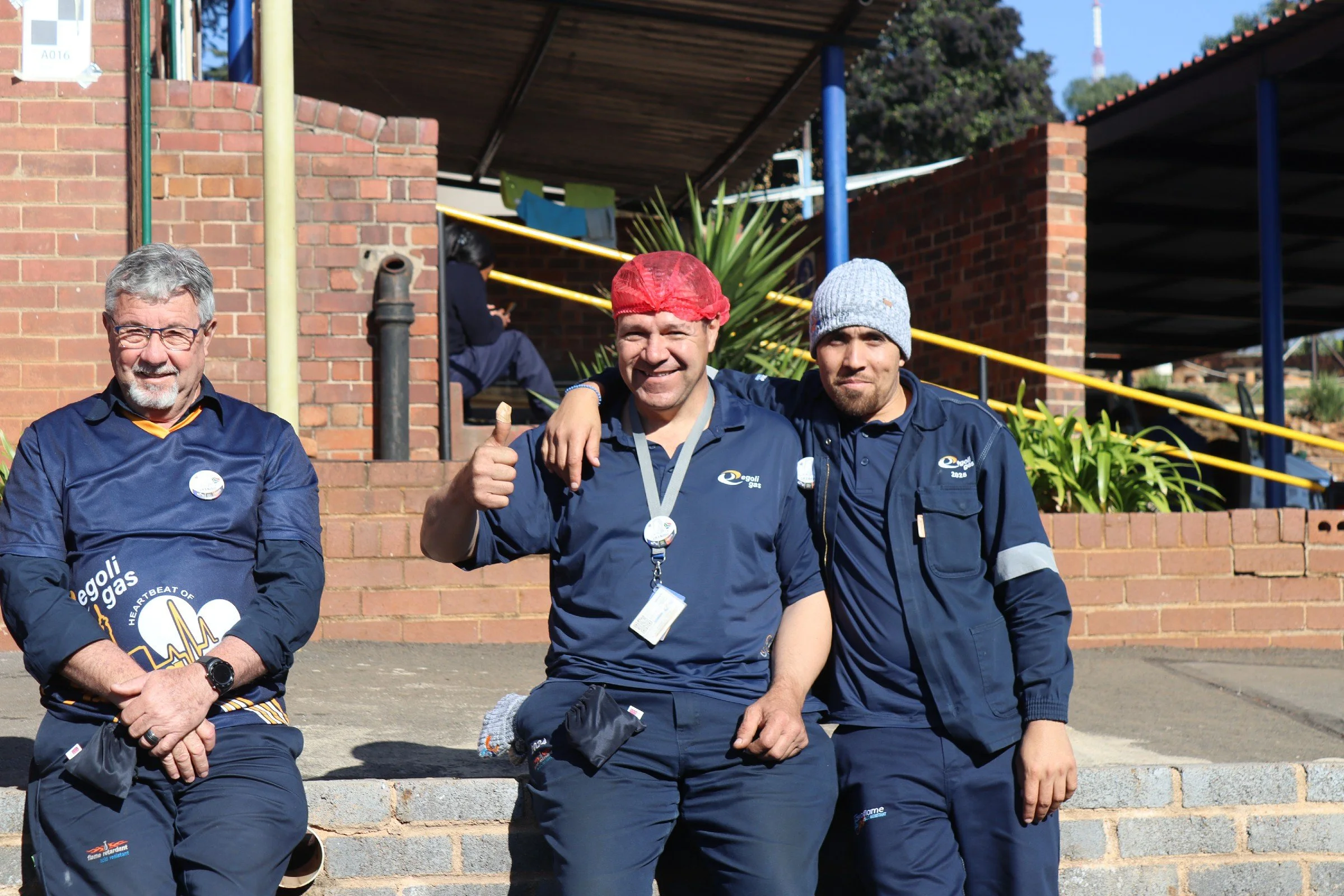 Four men wearing blue shirts, two with medals around their necks, posing outdoors on steps in front of a brick wall, with one man giving a thumbs-up. They appear to be celebrating or participating in an event.