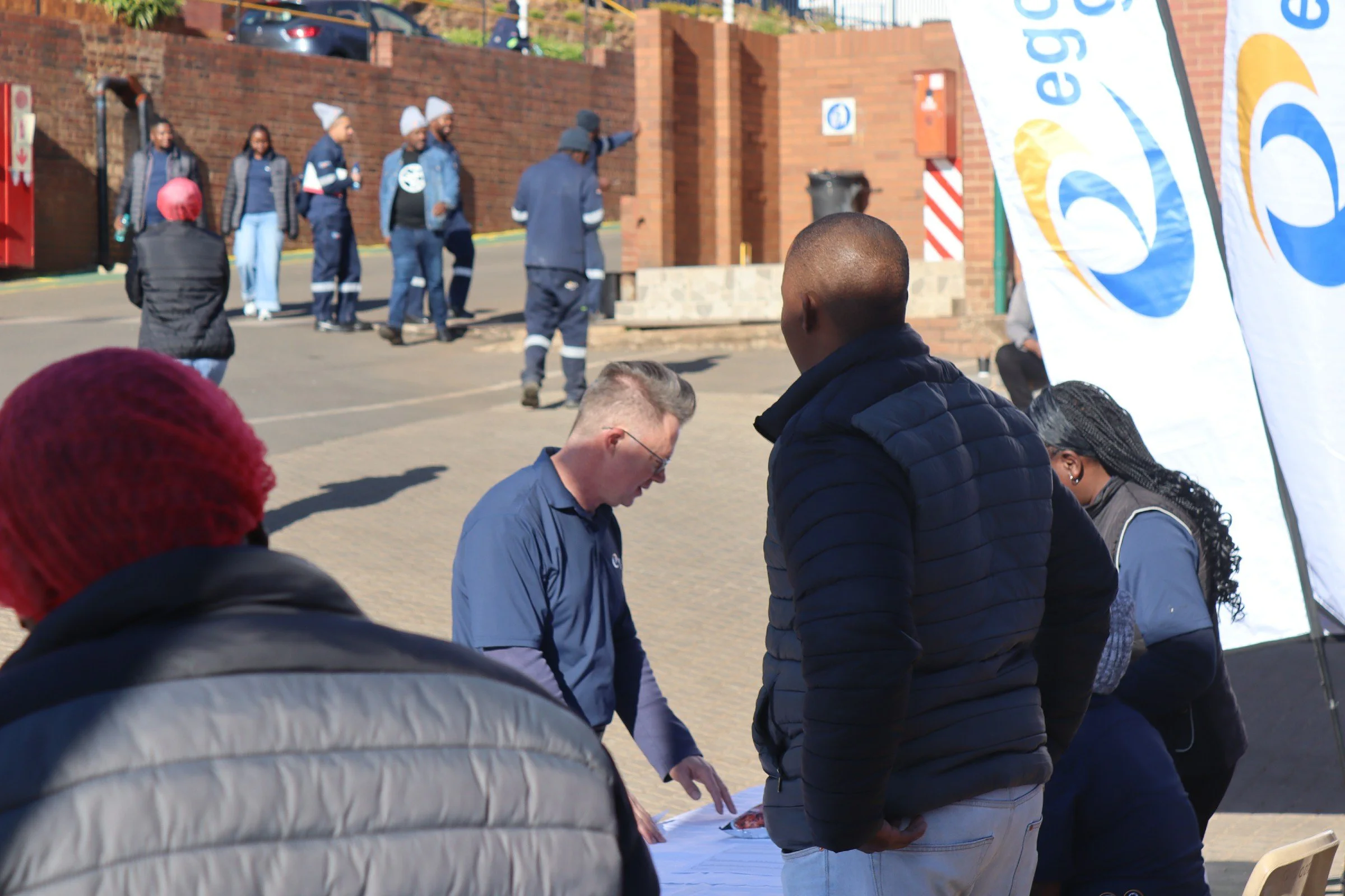 People standing in a line outdoors, possibly at an event or gathering, with a brick wall and posters in the background.