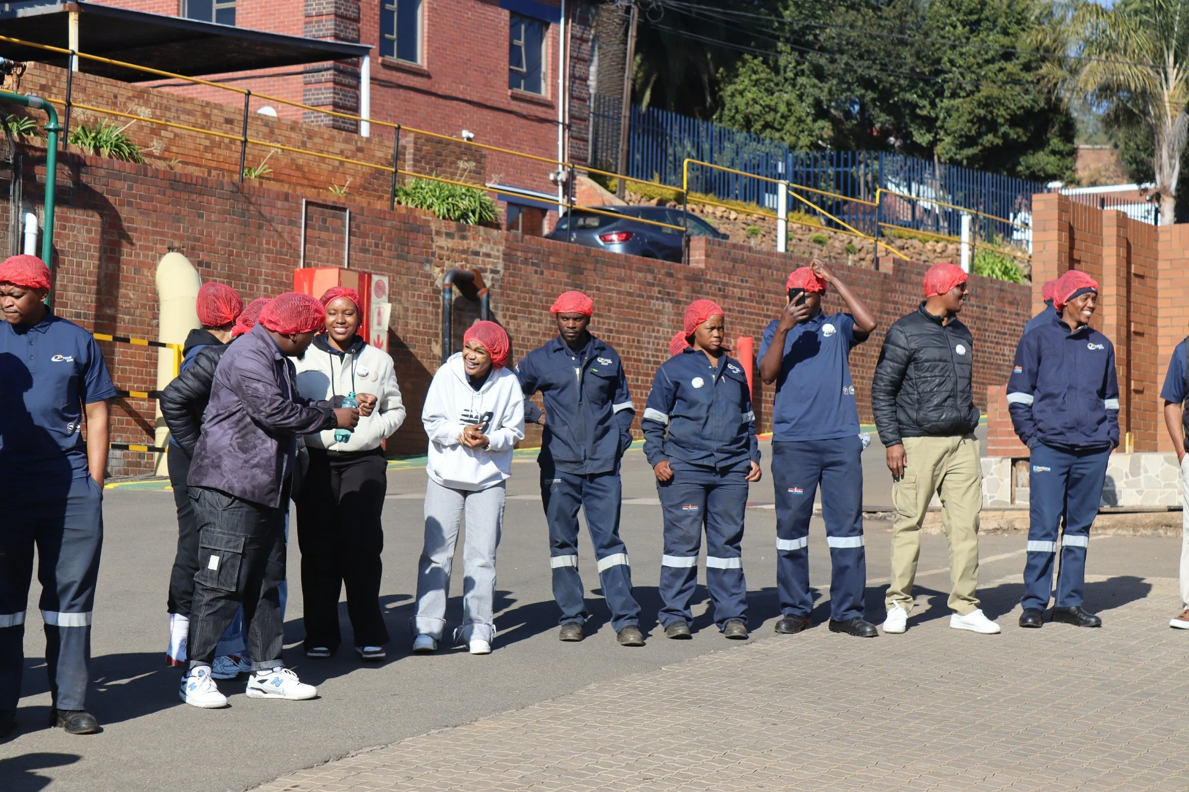 Group of people wearing red hairnets standing outside near a brick building, some smiling and talking, under a sunny sky.