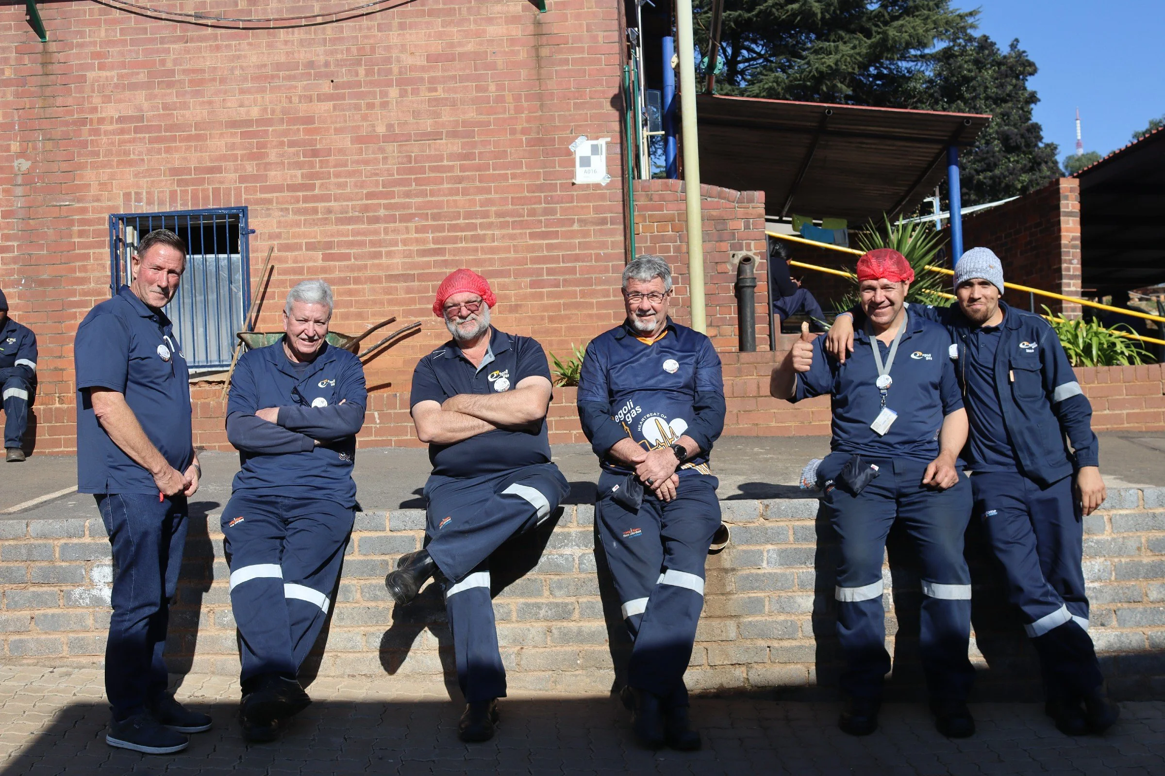 Group of seven men in navy uniforms posing outdoors on steps in front of a brick building, some with arms crossed, one giving a thumbs-up, and wearing red or gray beanies or hats.