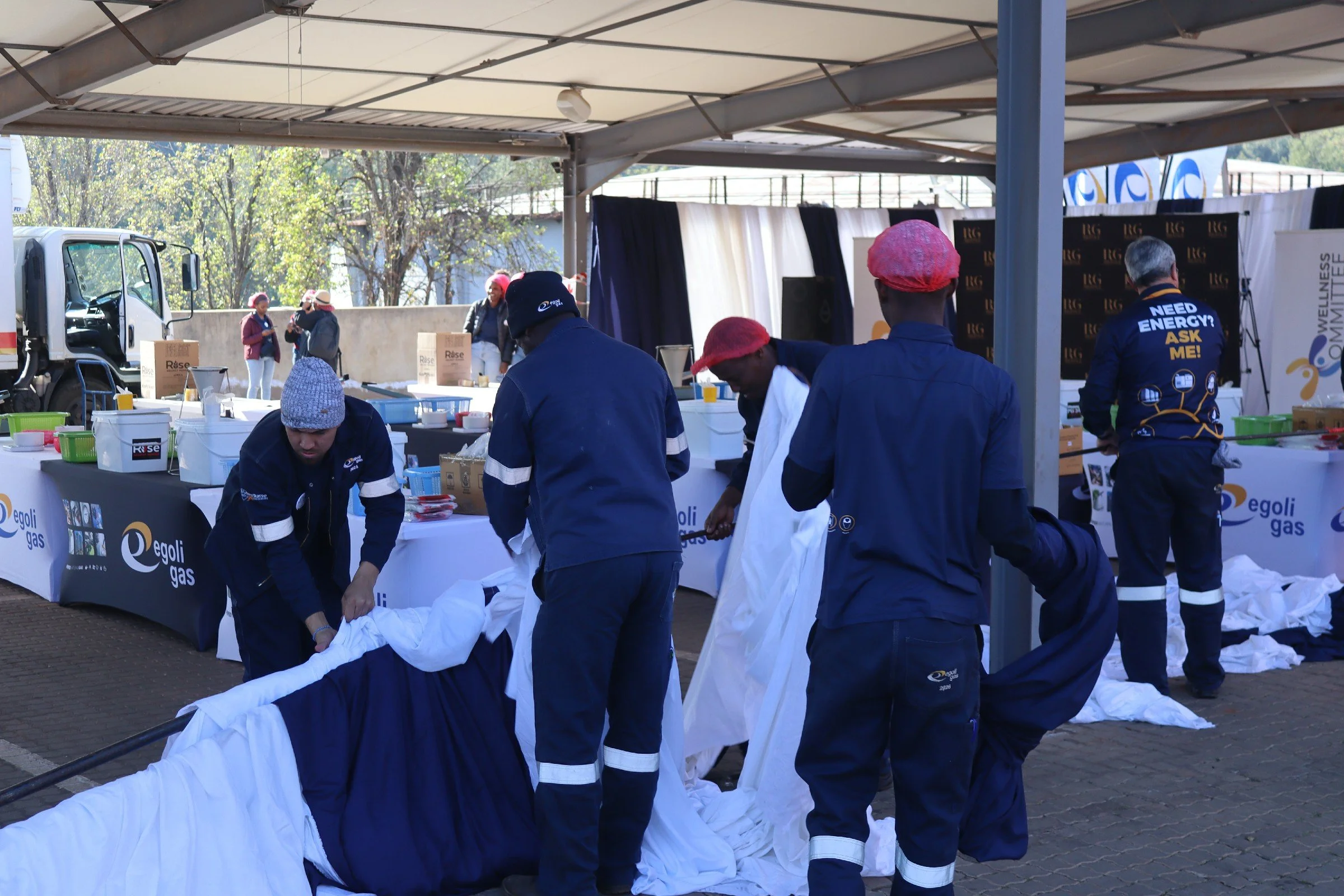 Emergency personnel wrapping a body in a white cloth at an outdoor event under a canopy, with tables and banners in the background.
