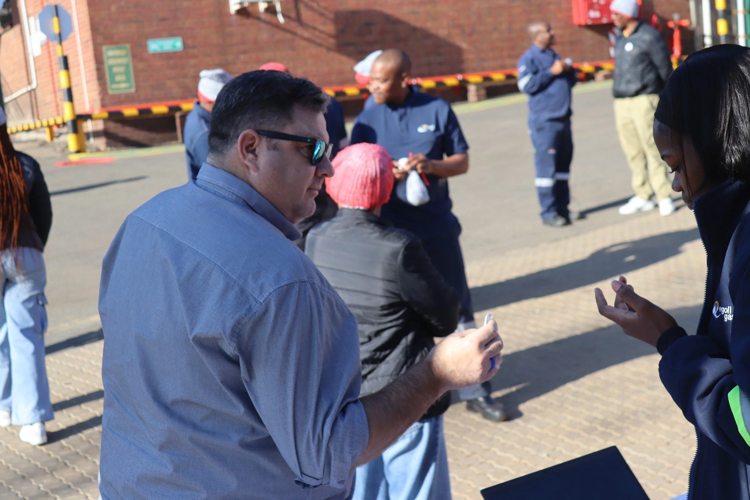 Group of people standing outside on a brick-paved area, some talking and others waiting, with a brick building and a street sign in the background.