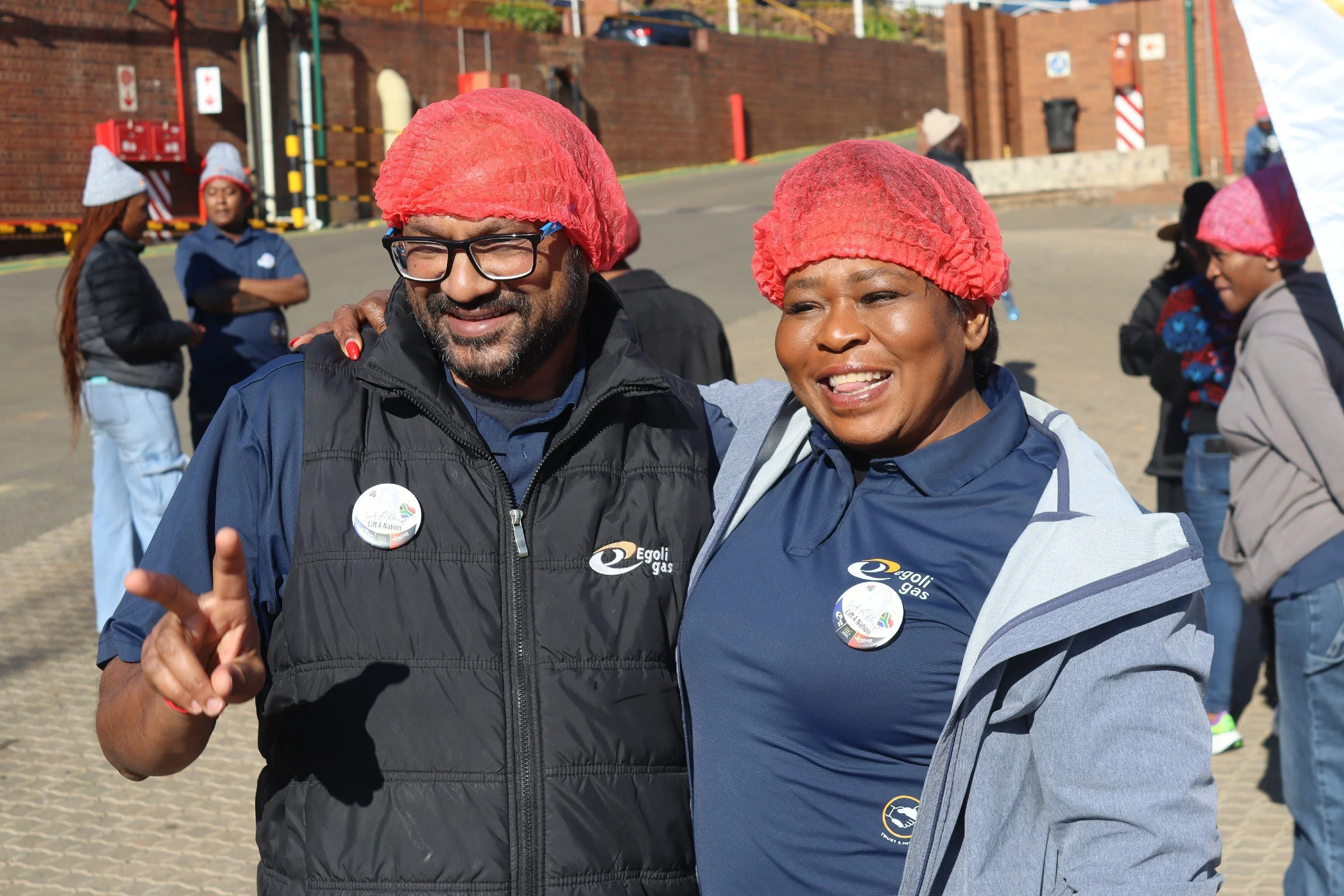 Two smiling people with red hairnets posing outdoors, with several other people in the background. The man on the left is wearing glasses and a black vest with the logo 'Egolig Gas,' and the woman on the right is wearing a blue jacket. They are stand