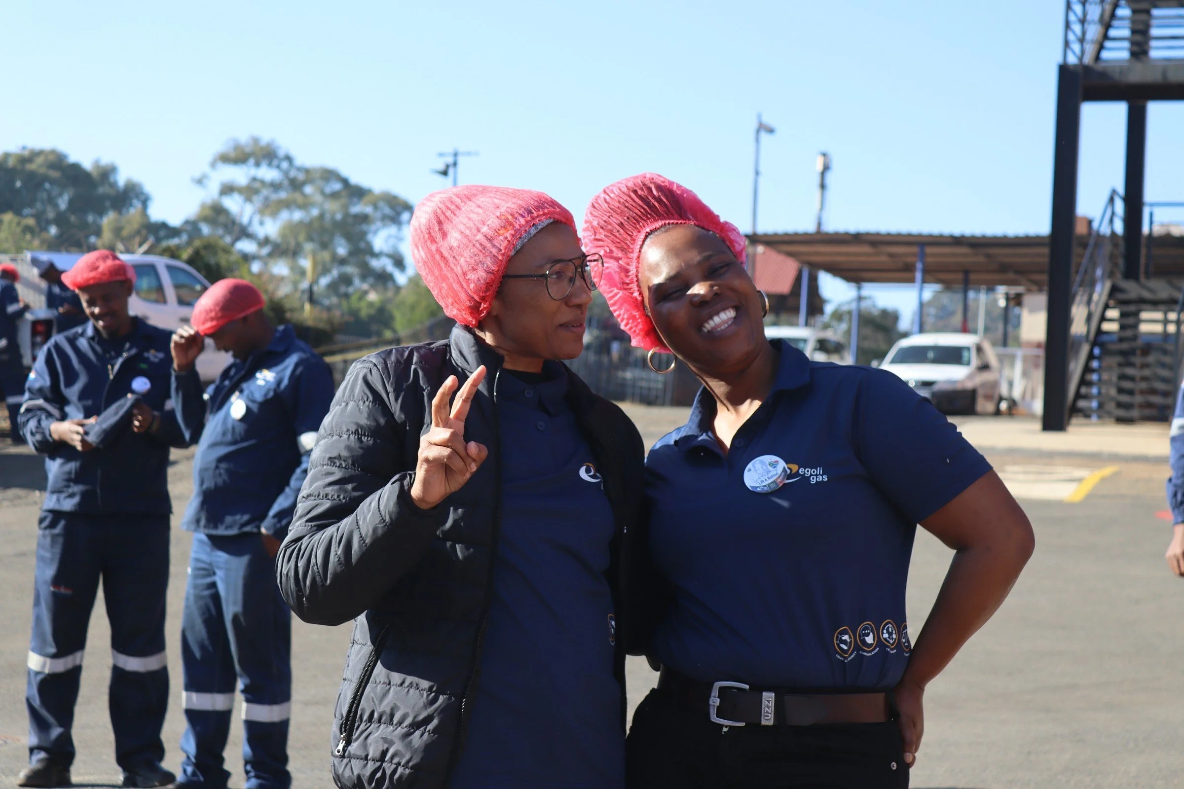 Two women, wearing pink shower caps and navy shirts with logos, are smiling and taking a selfie together. In the background, there are several people and vehicles, suggesting an outdoor event or gathering, possibly related to energy or utilities.
