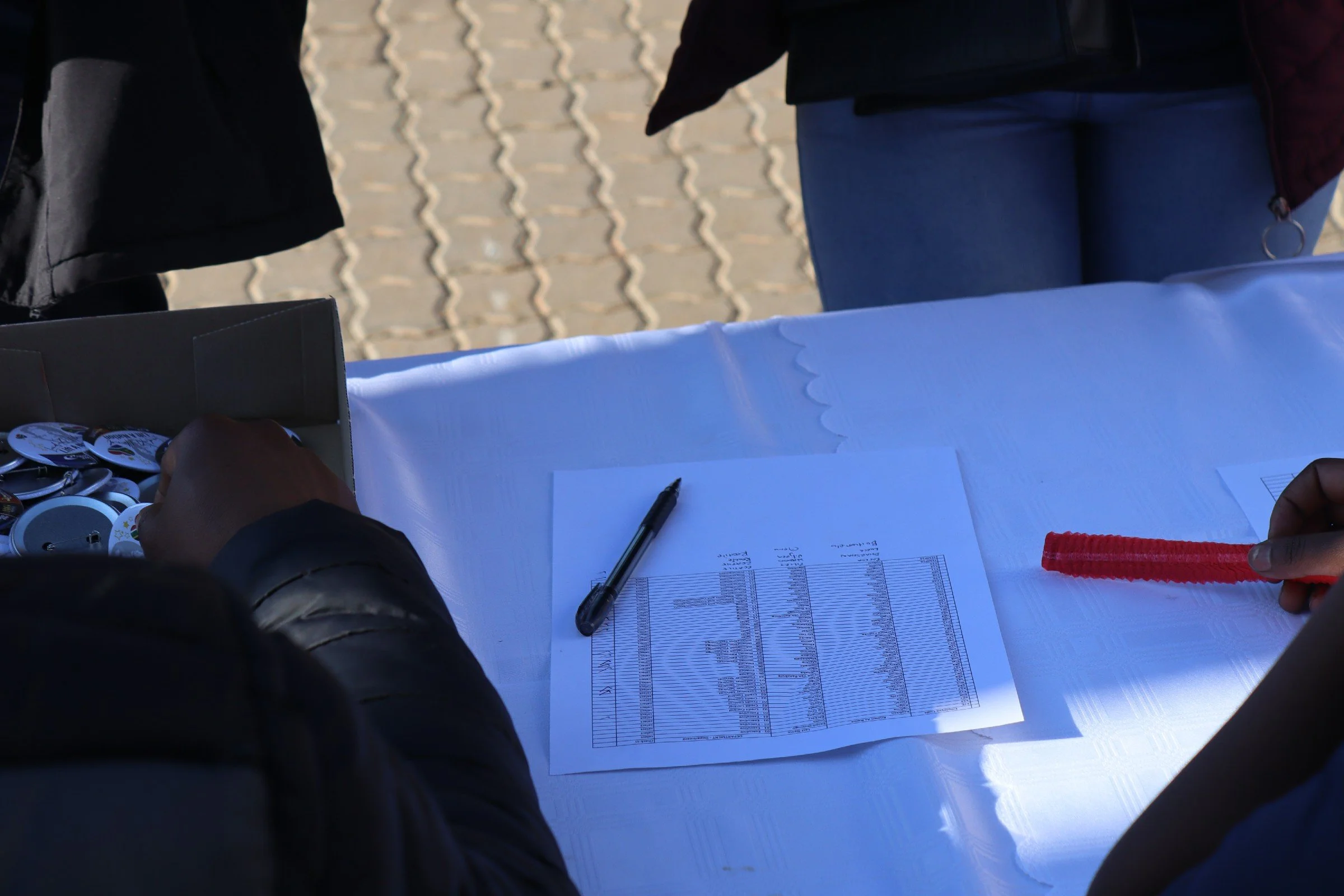 People at a table with papers and a pen, outside on a brick-paved area.