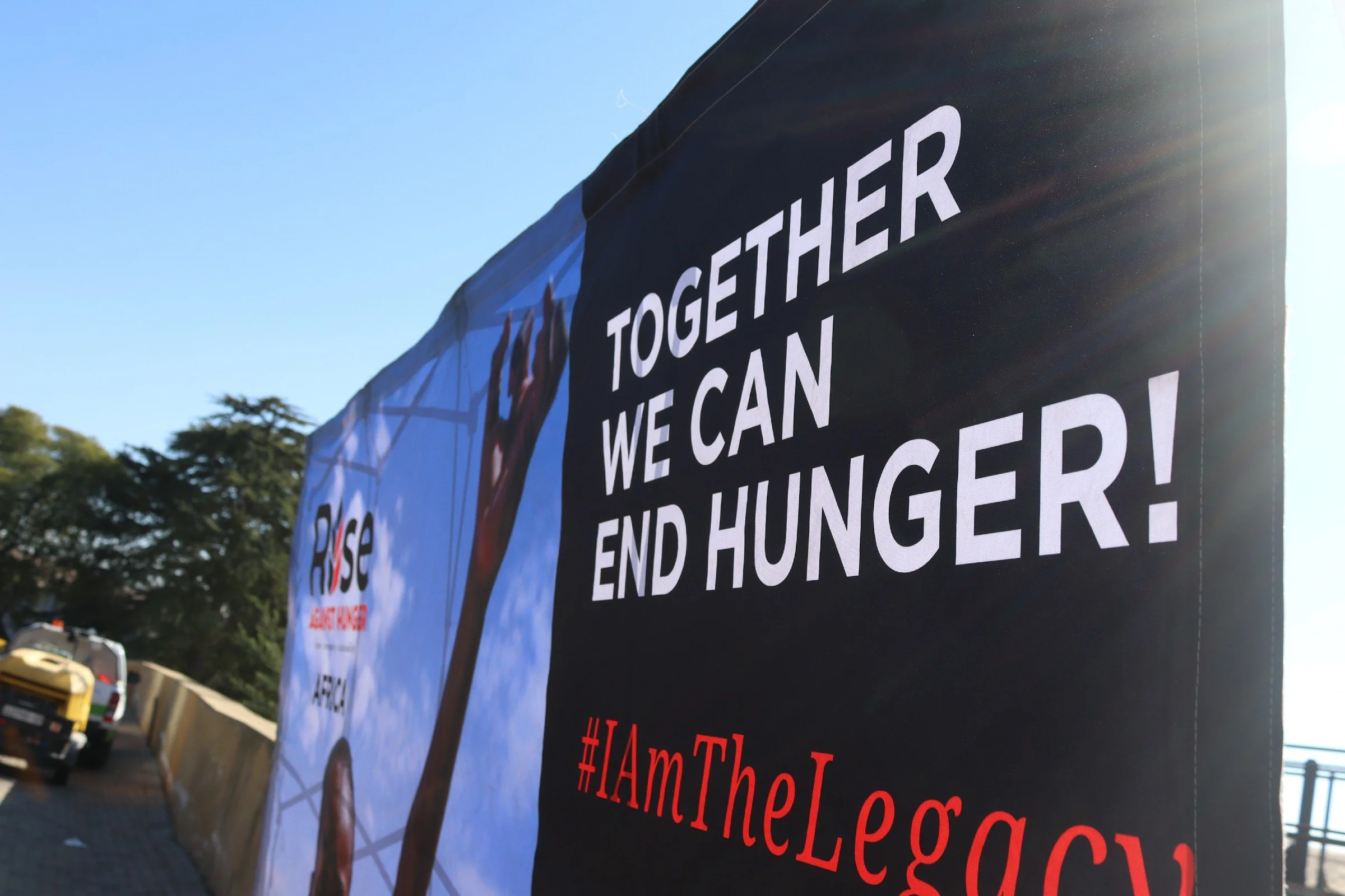 A protest banner with the message 'Together we can end hunger!' is displayed outdoors, with trees, vehicles, and a clear sky in the background.