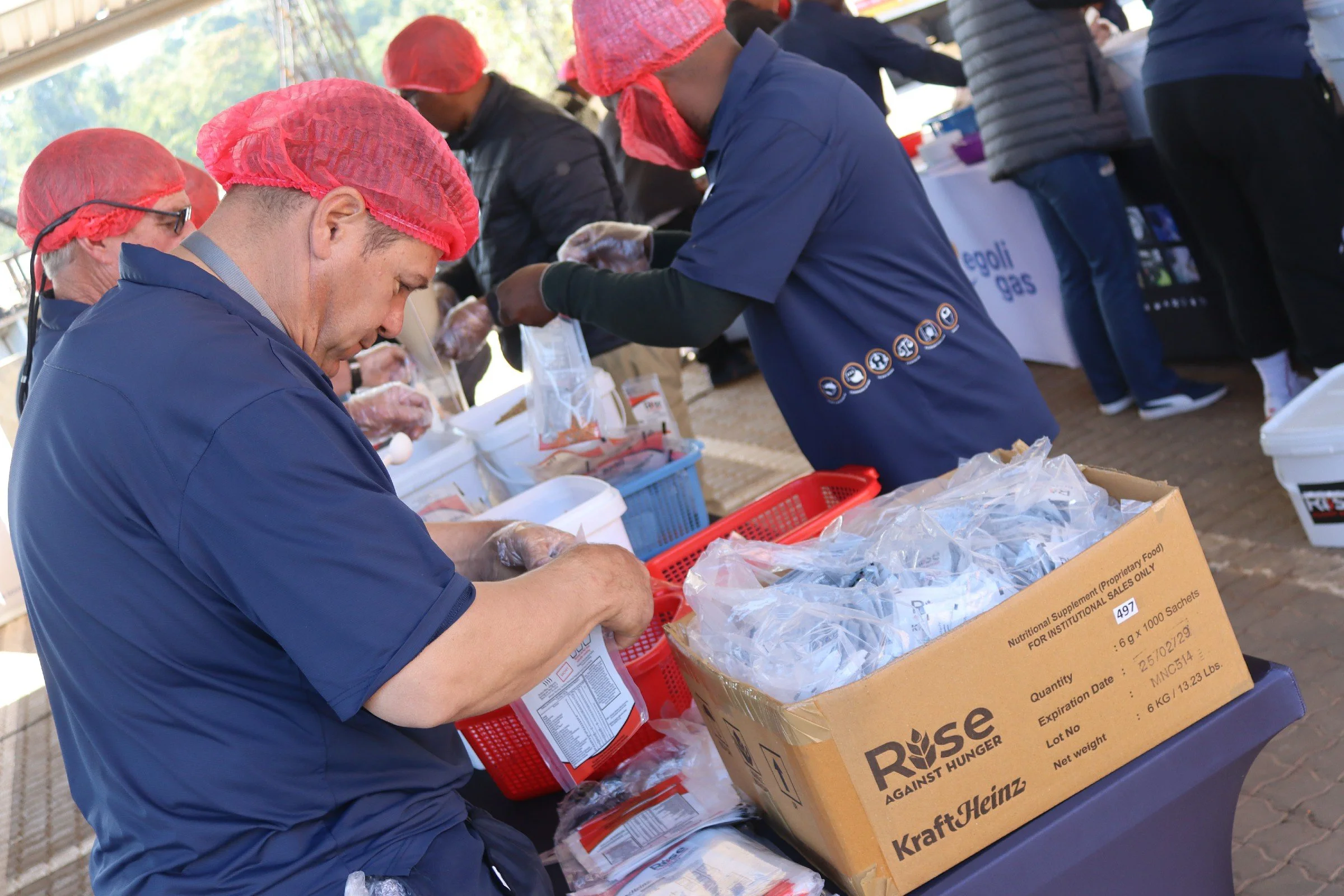 People wearing red hairnets and gloves, sorting and packaging food items at a table outdoors during a food drive or charity event.