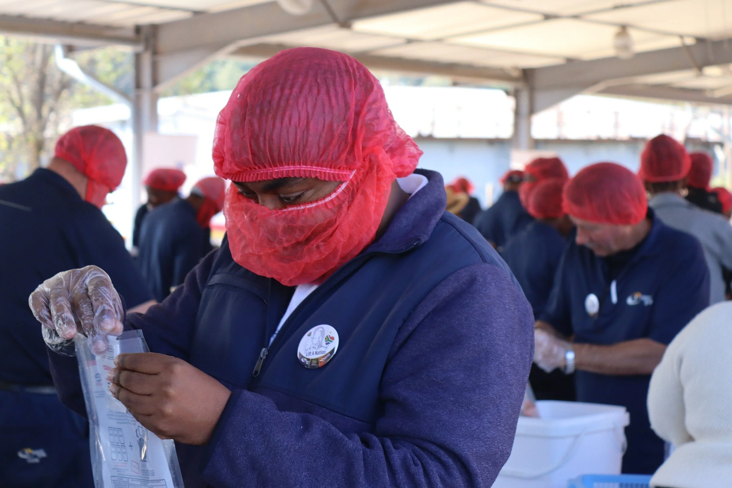 A group of people, wearing red hair coverings and dark jackets, working together under a sheltered outdoor area, possibly packaging or preparing items.
