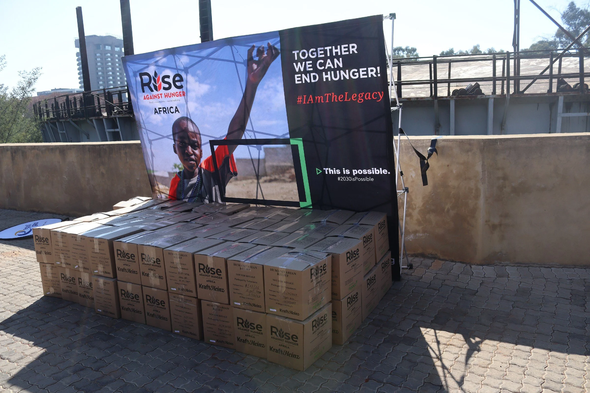 Banners promoting a hunger relief event called Rise Against Hunger Africa with a message about ending hunger and a hashtag #IAmTheLegacy. Several boxes labeled 'Kraft Heinz' and 'Rise Against Hunger' are stacked on the ground.