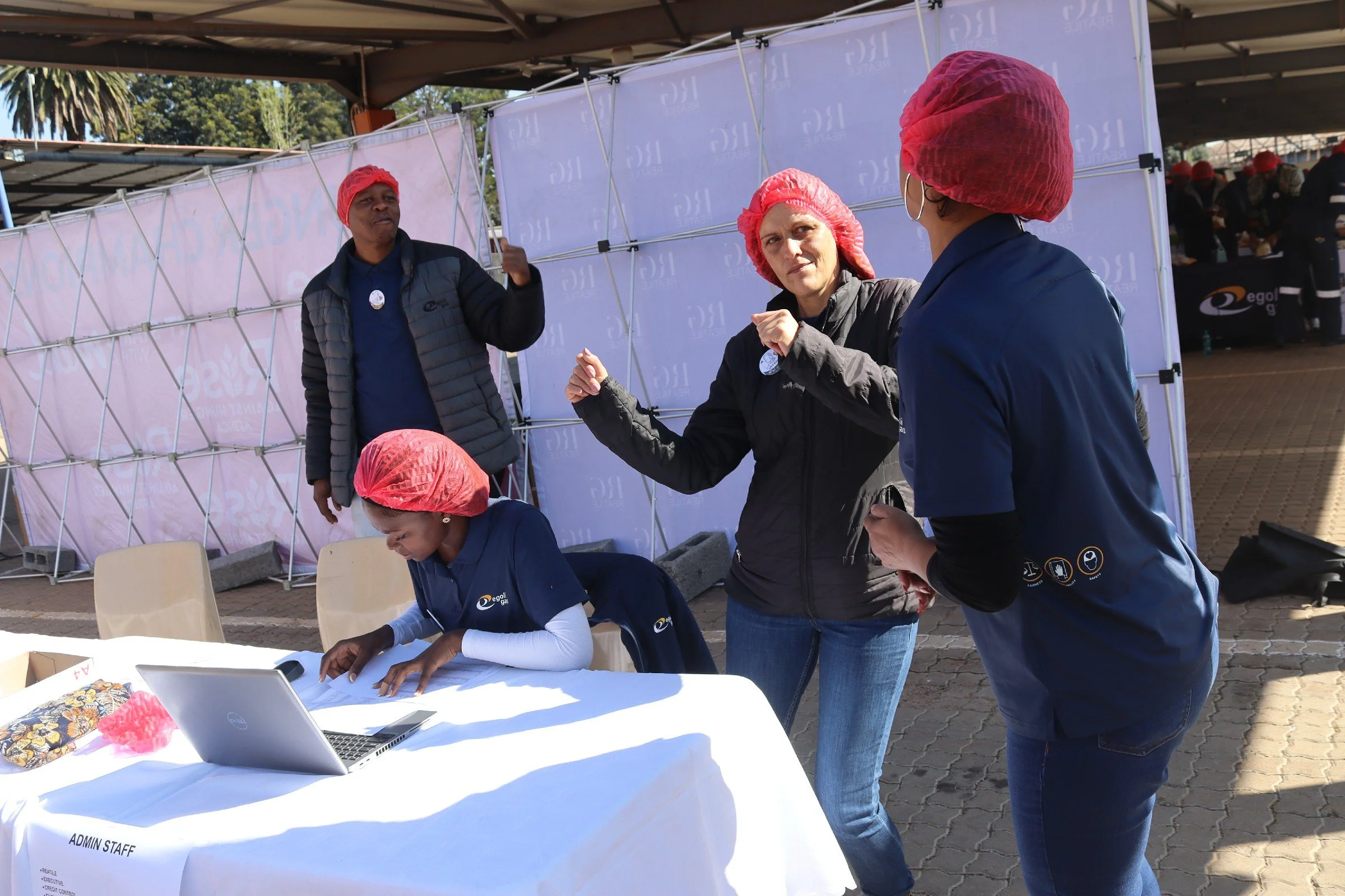 People working at an outdoor event booth with laptops, wearing pink hair covers, under a canopy with a purple backdrop.