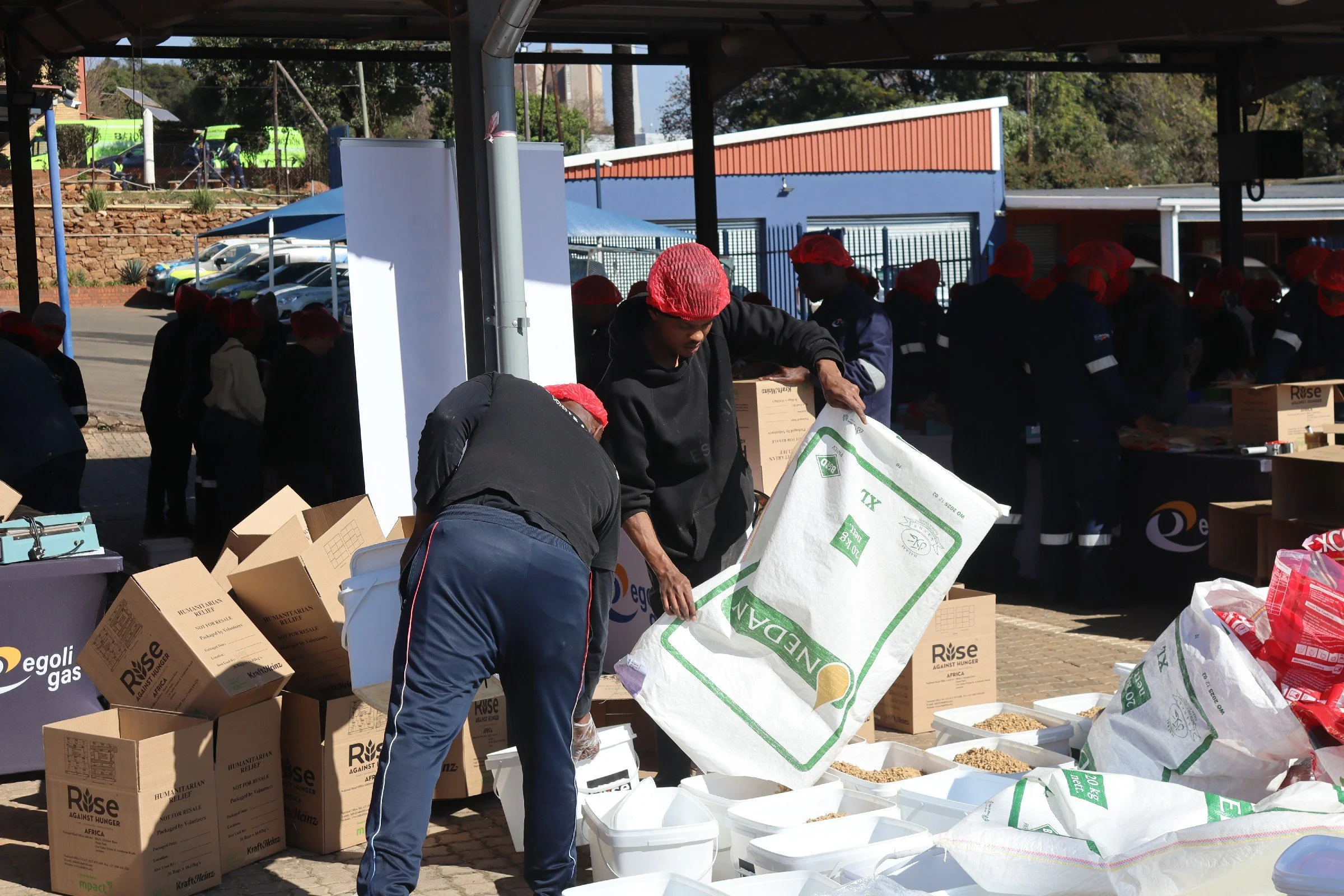 People wearing red hair nets packing food into boxes at a charity event with boxes and bags on tables and the background showing parked cars, a building, and trees.