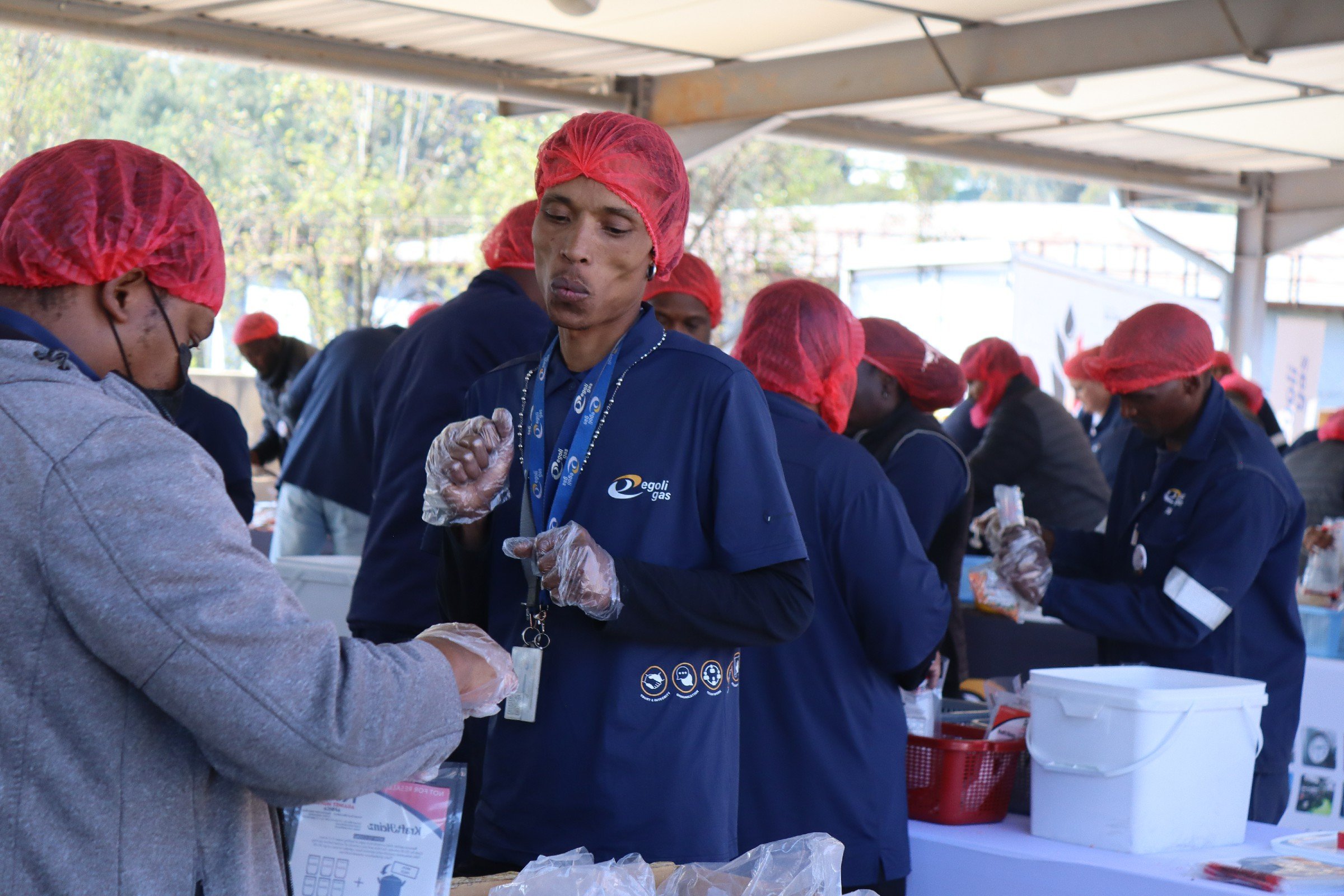People working at a community service event, wearing red hairnets and gloves, under a tent with trees in the background.