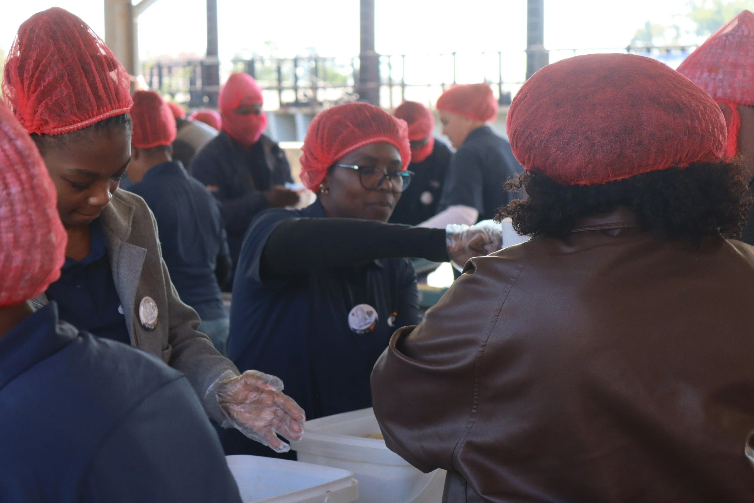 People wearing red hairnets and gloves working together in a food processing or assembly setting, with a bright background of large windows and outdoor scenery.