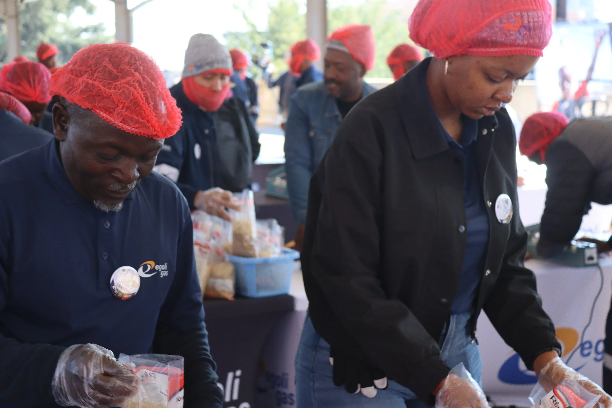 People working together at a community event, some wearing red hairnets and gloves, packing food items on tables under a pavilion with trees in the background.