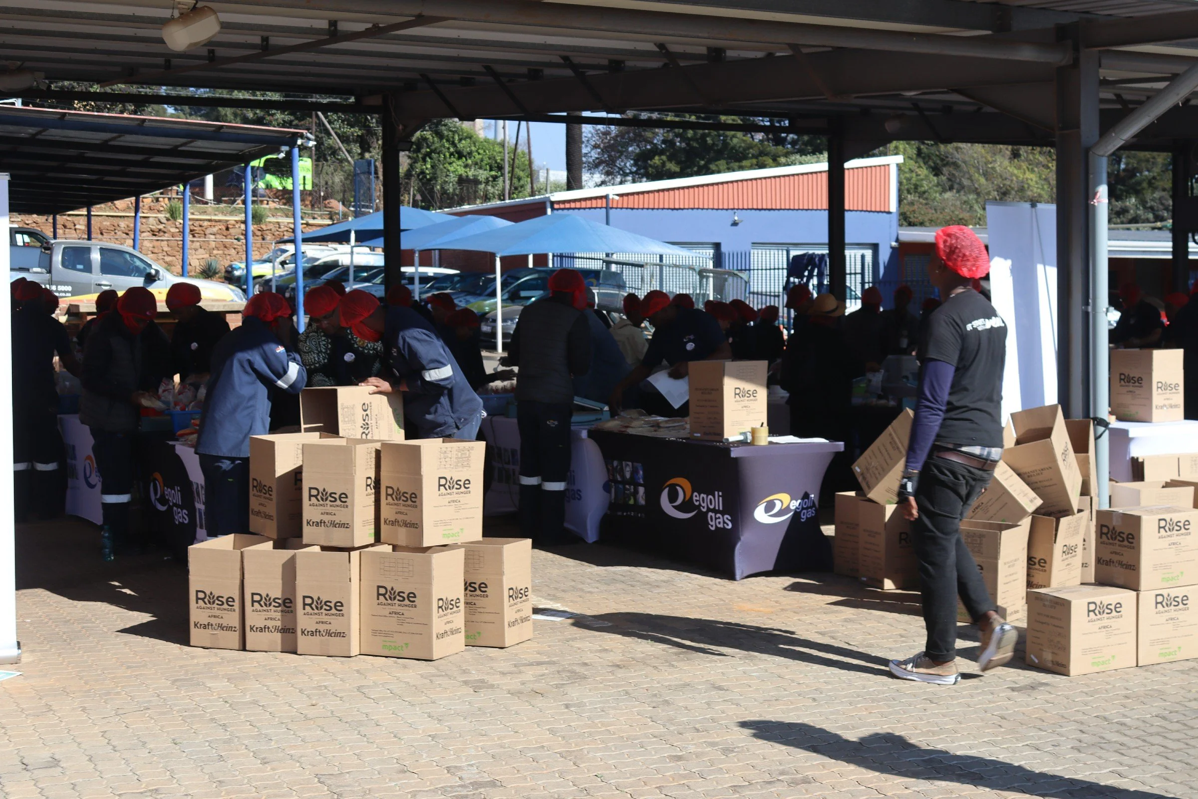 People wearing red head coverings working at tables under a covered outdoor area, with stacked boxes labeled 'Rise Against Hunger' and other people visible in the background.