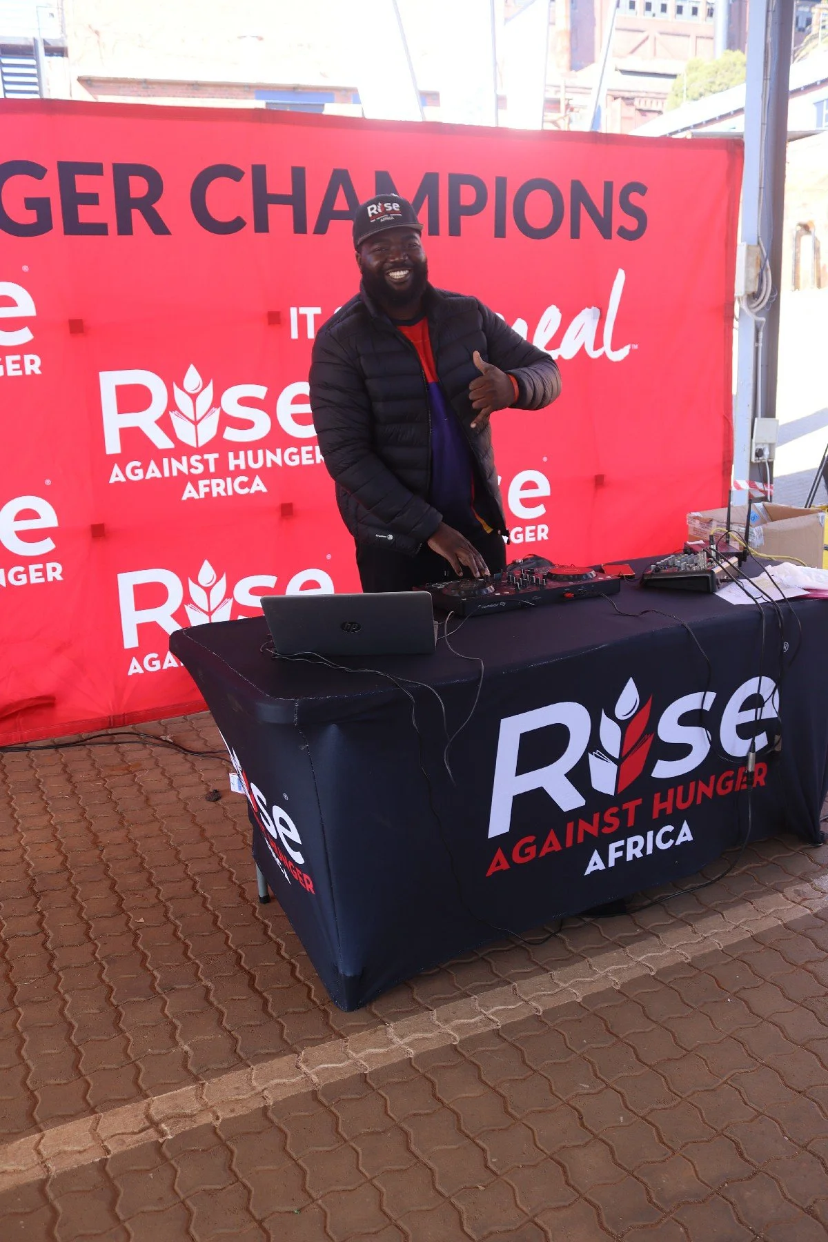 A man smiling and giving a thumbs up at a DJ setup during an event for Rise Against Hunger Africa. He is standing behind a black table with electronic equipment, in front of a red backdrop with Rise Against Hunger Africa branding.