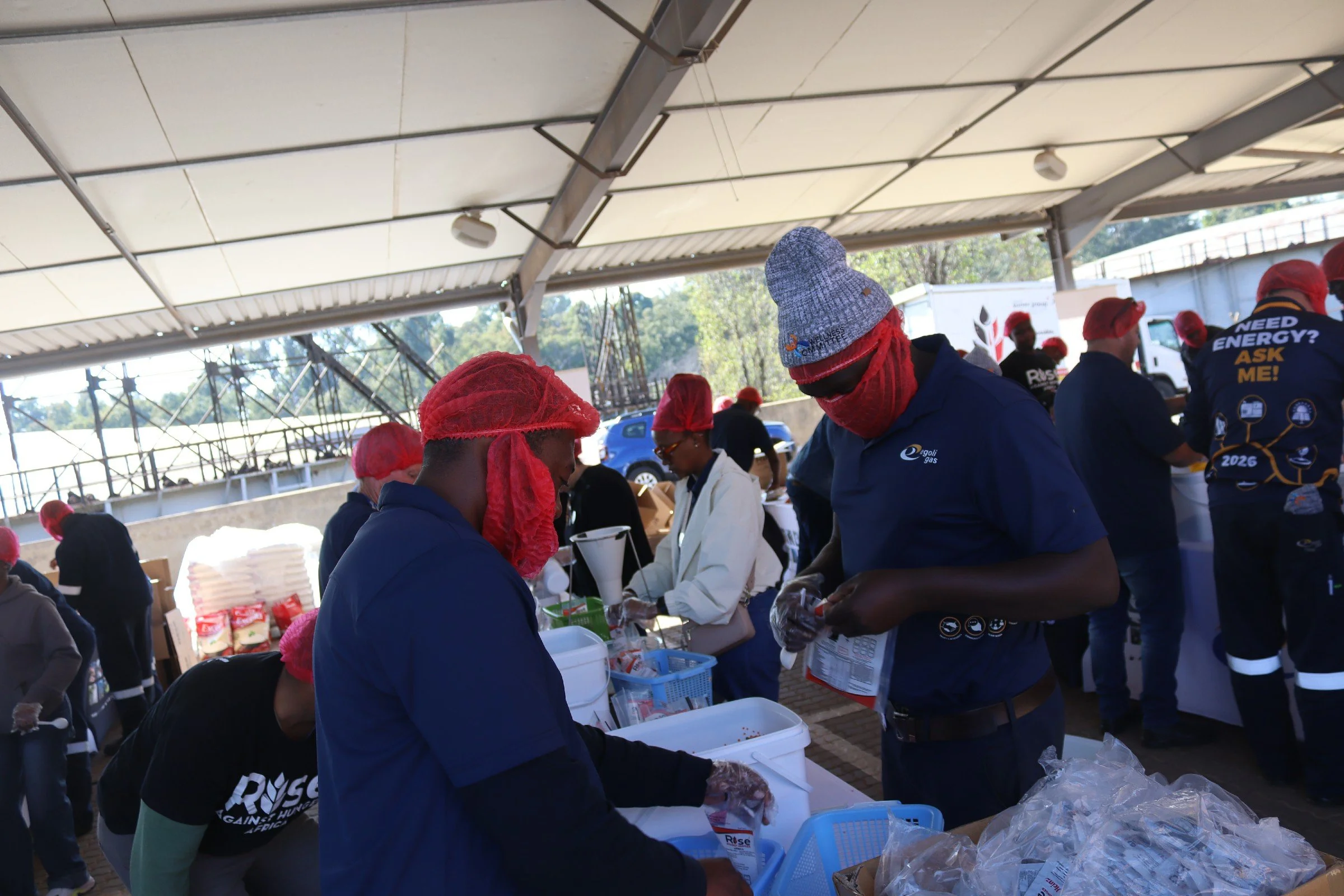 People working at tables under a large outdoor shelter, wearing red hairnets, with some in masks, and stacks of supplies or food items on the tables.