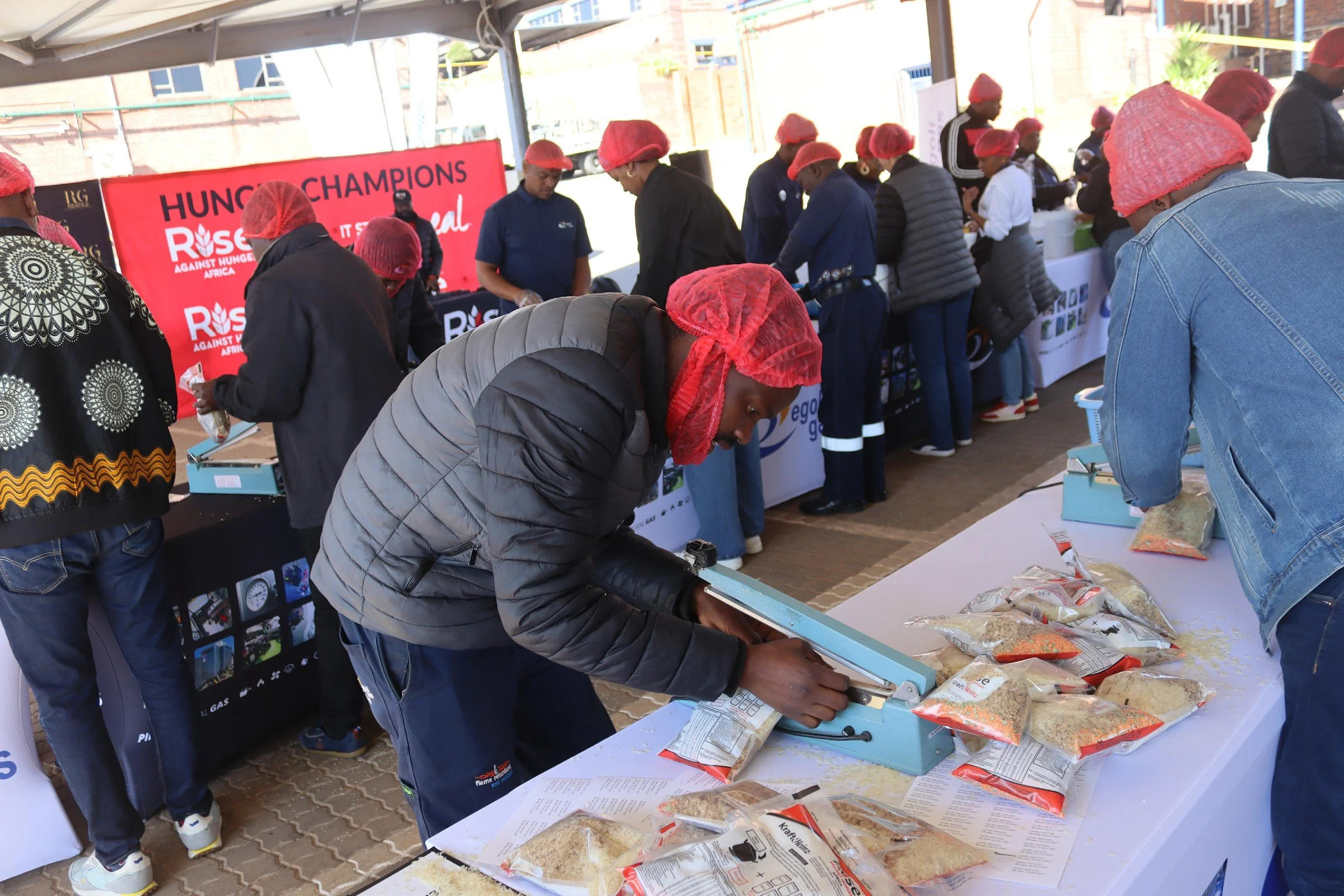 People wearing red hairnets at a food distribution event, with a red banner in the background that reads "HUNGER CHAMPIONS RISE AGAINST HUNGER AFRICA," and tables with food packages and scales for packing food.