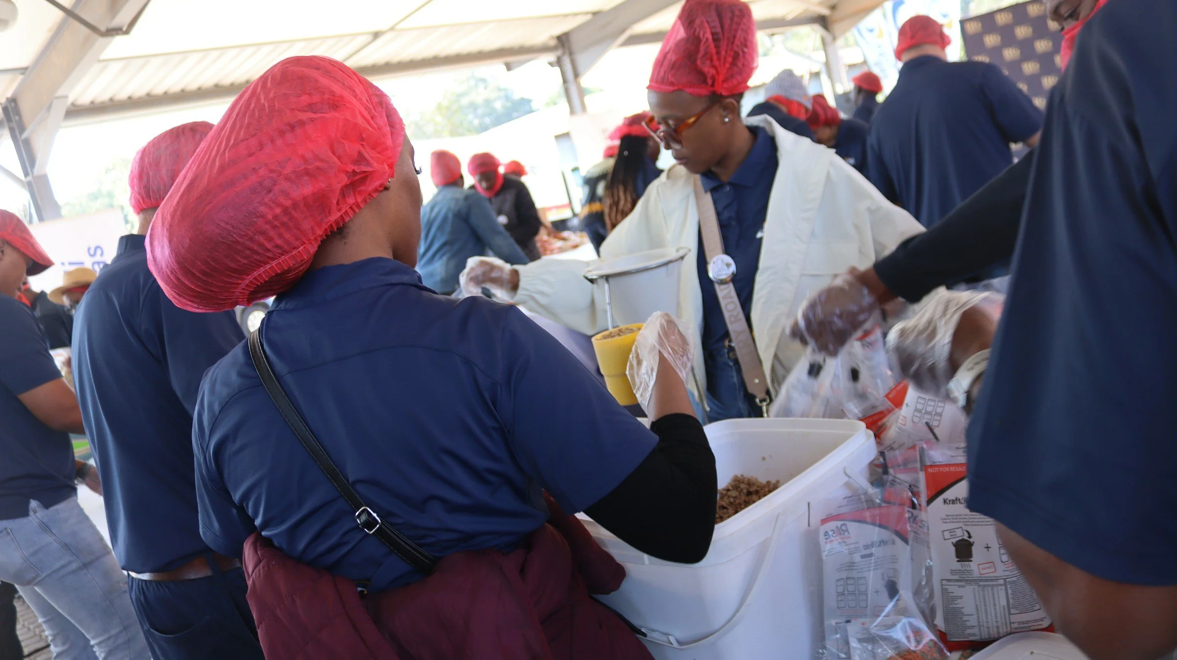 People wearing red hairnets and gloves preparing and serving food at a community event, while others are working in the background.