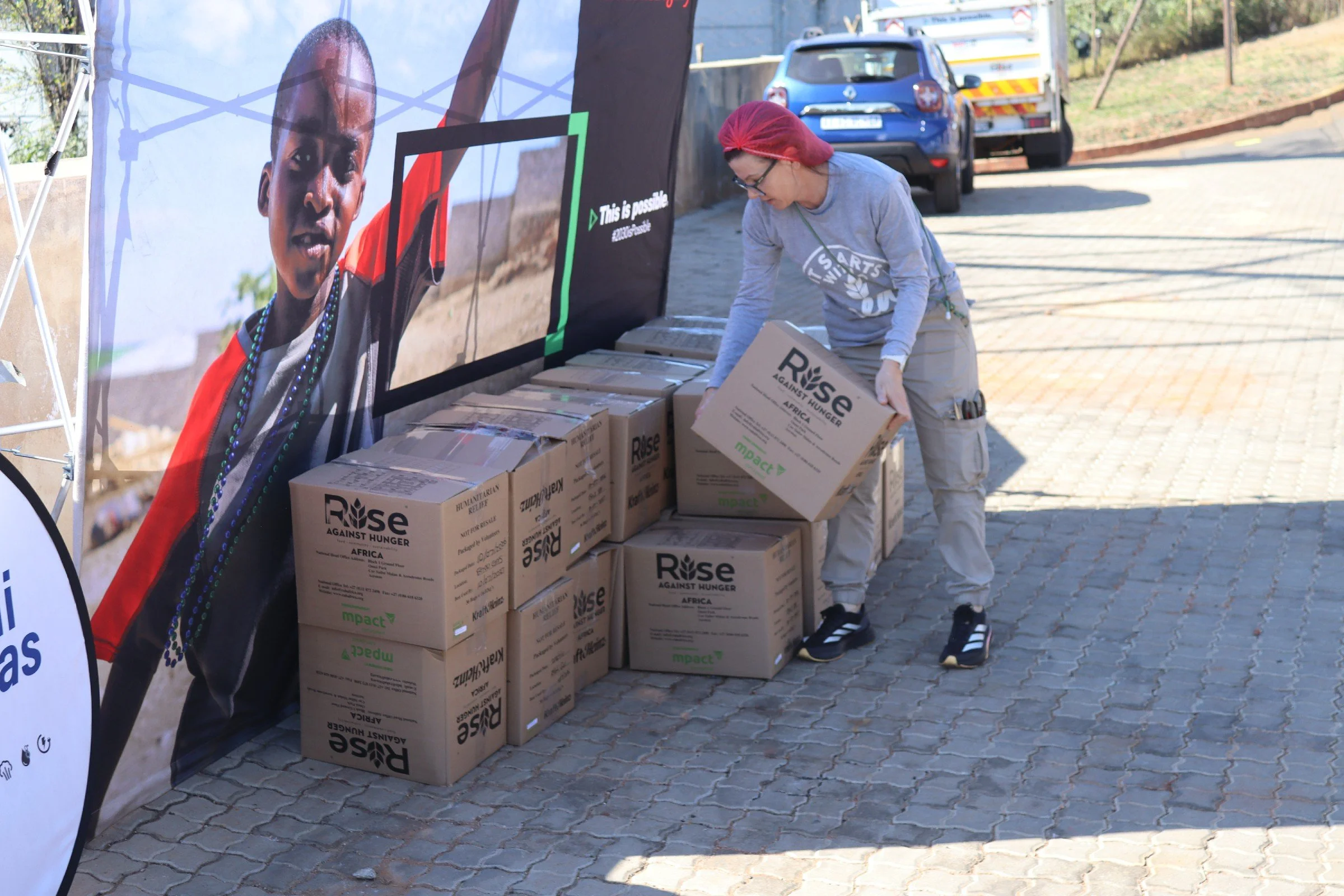 A woman wearing glasses, a gray long-sleeve shirt, and a red headscarf sorts boxes labeled 'Rise Against Hunger' at an outdoor event.