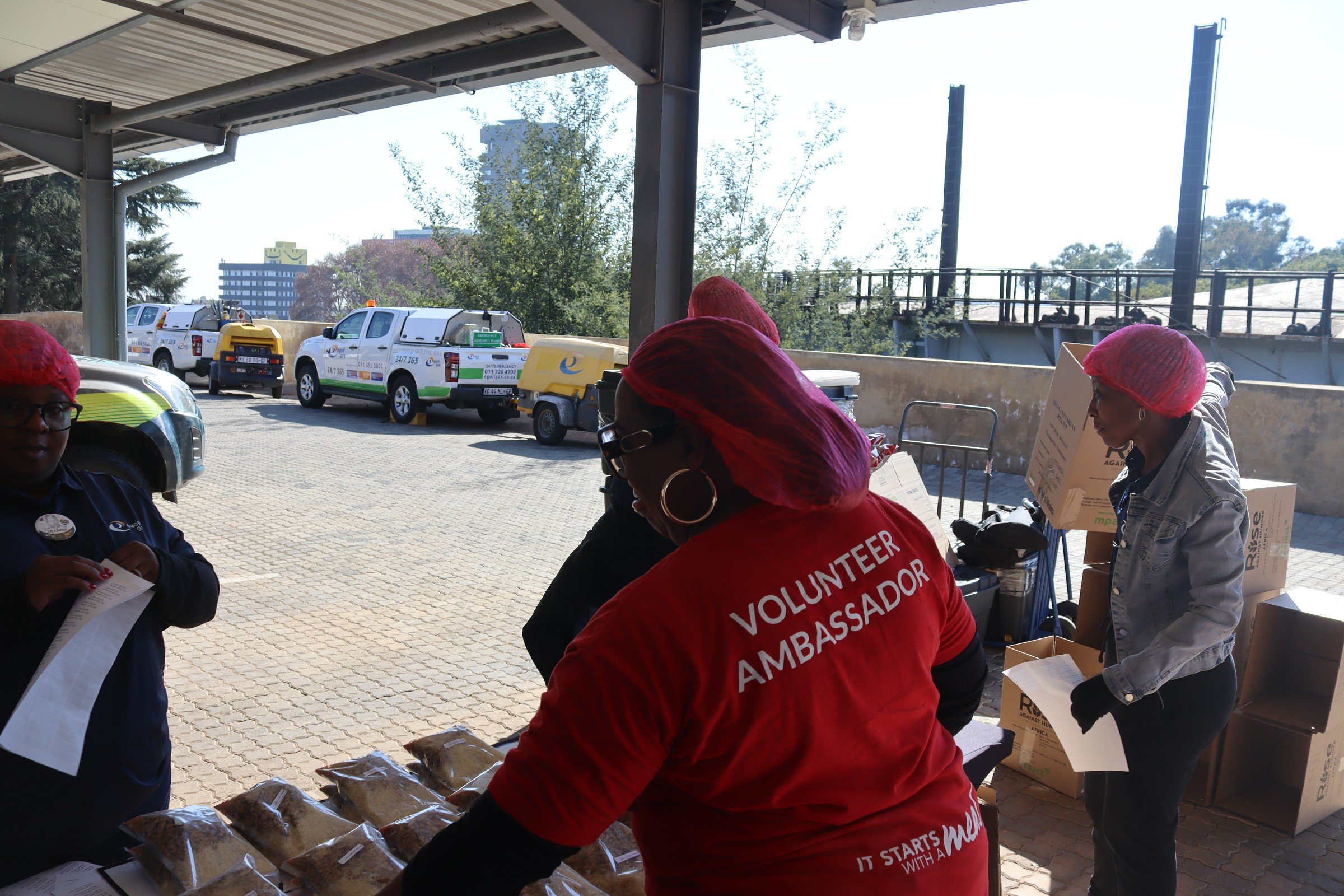 People wearing red volunteer vests and hair coverings are gathered at an outdoor table with food items, under a shaded area with parked vehicles and greenery in the background.