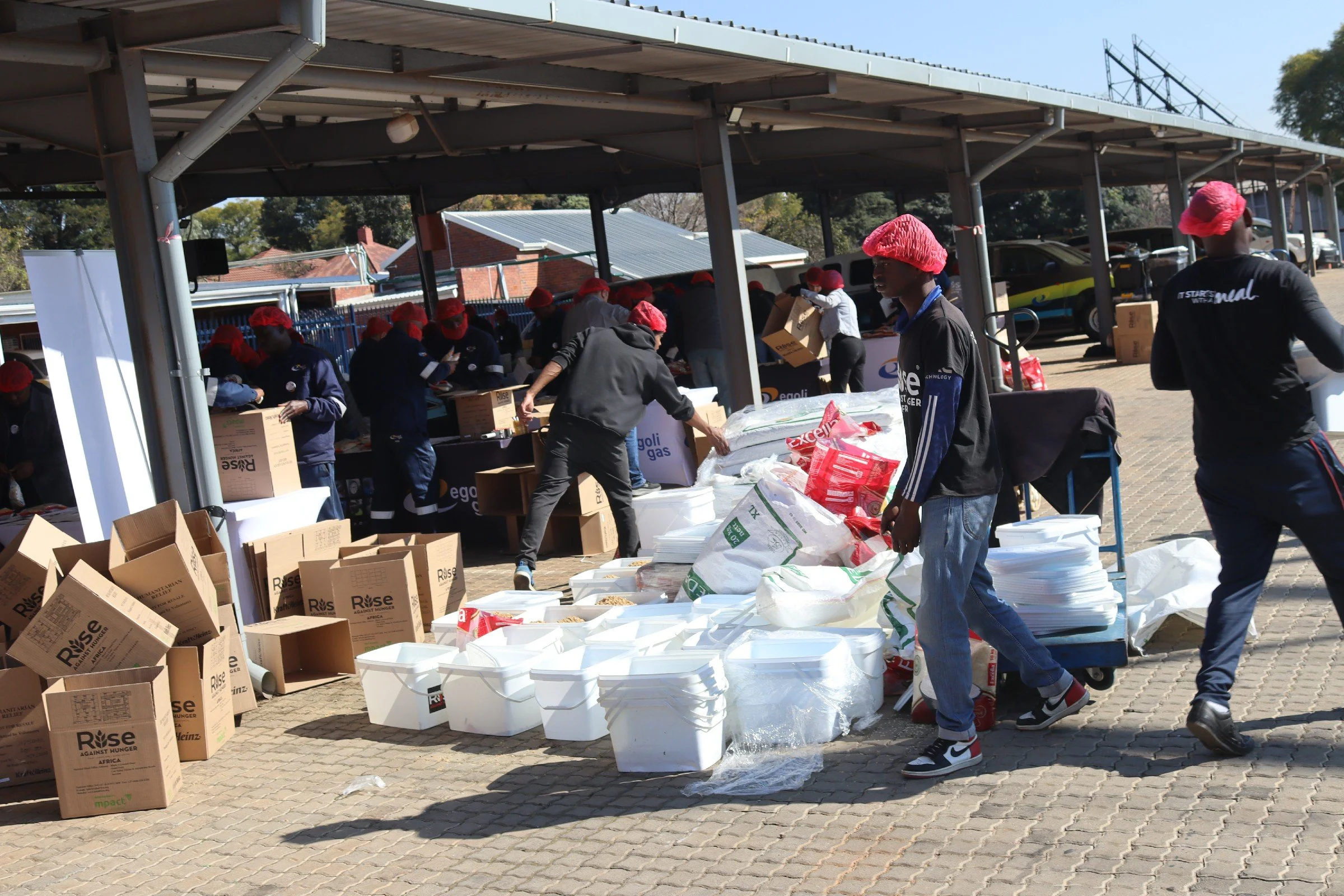 People wearing red hairnets and dark clothing are working outdoors at a long service table, packing boxes with supplies. Stacks of white containers and cardboard boxes labeled "Rise Against Hunger" are nearby. There are cars and a building in the bac