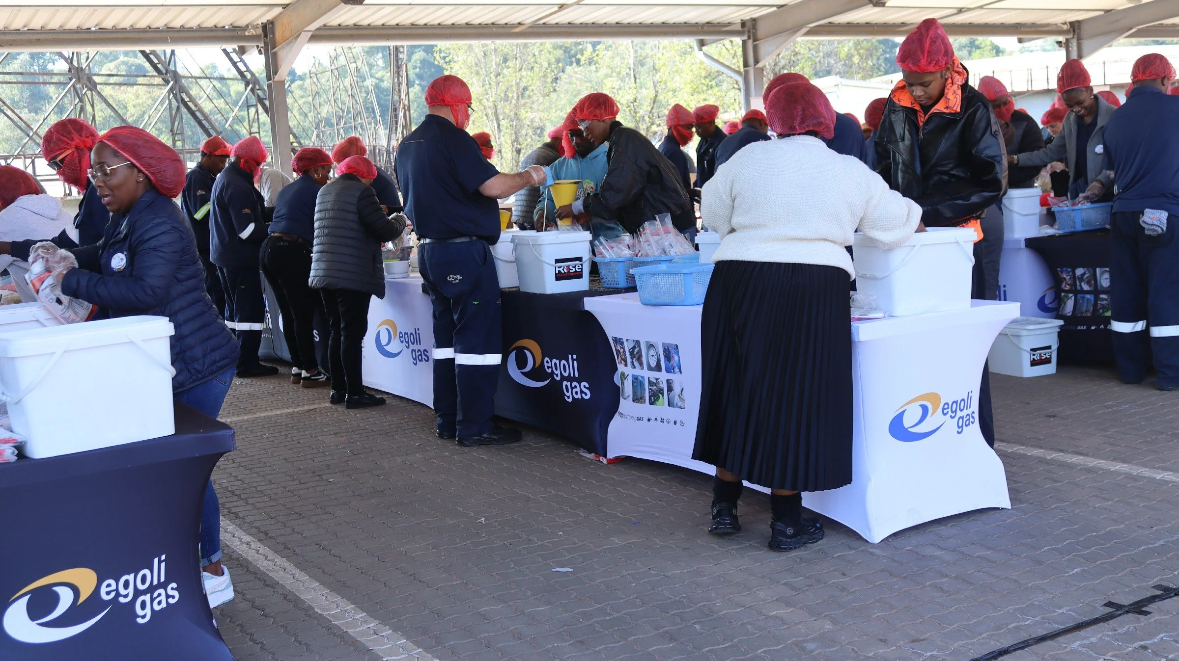 People wearing red hairnets and gloves at a food distribution event with tables branded with Egoli Gas logos under a covered outdoor area.