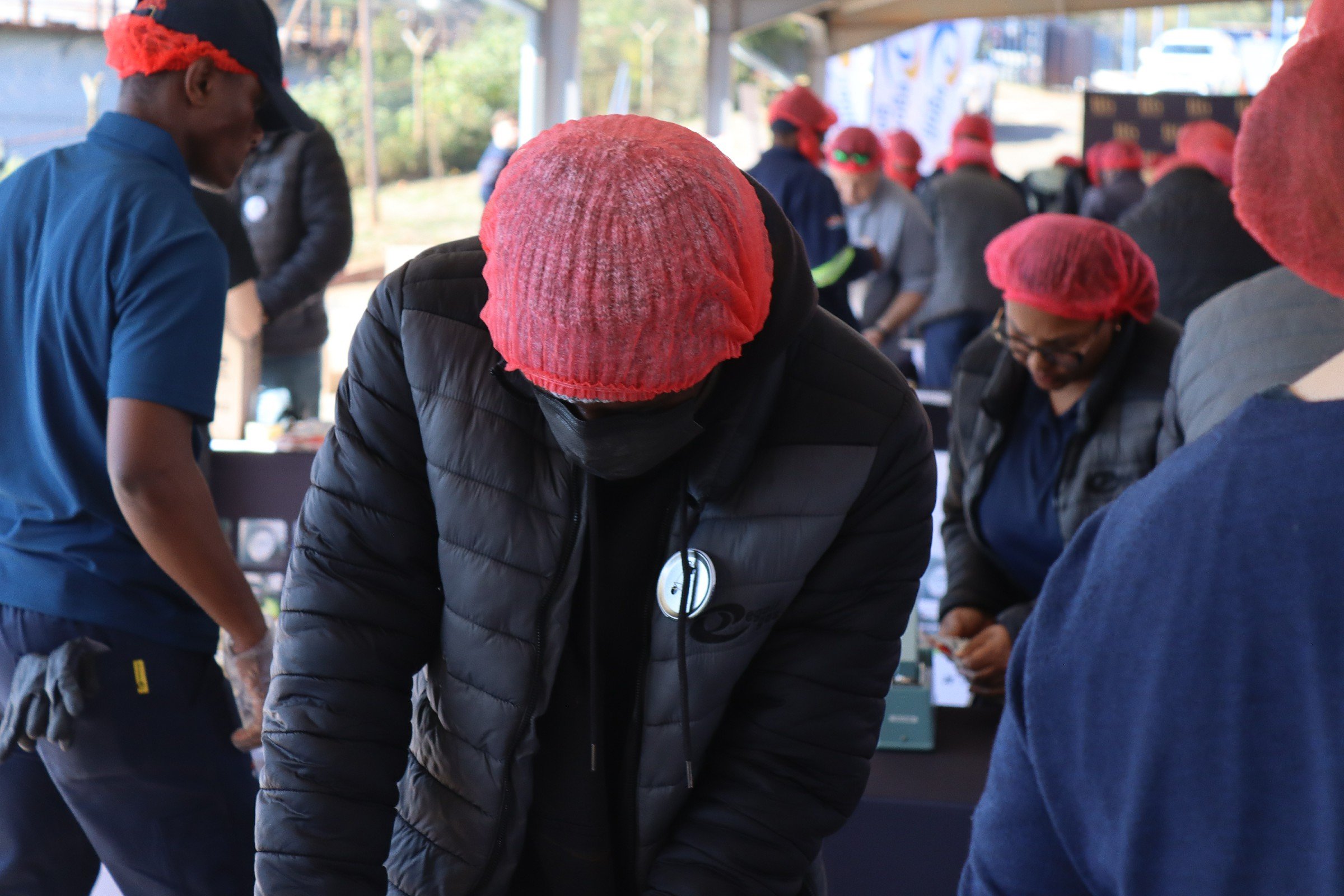 A group of people gathered outdoors, some wearing red headscarves and black masks, likely participating in a community event or service activity.