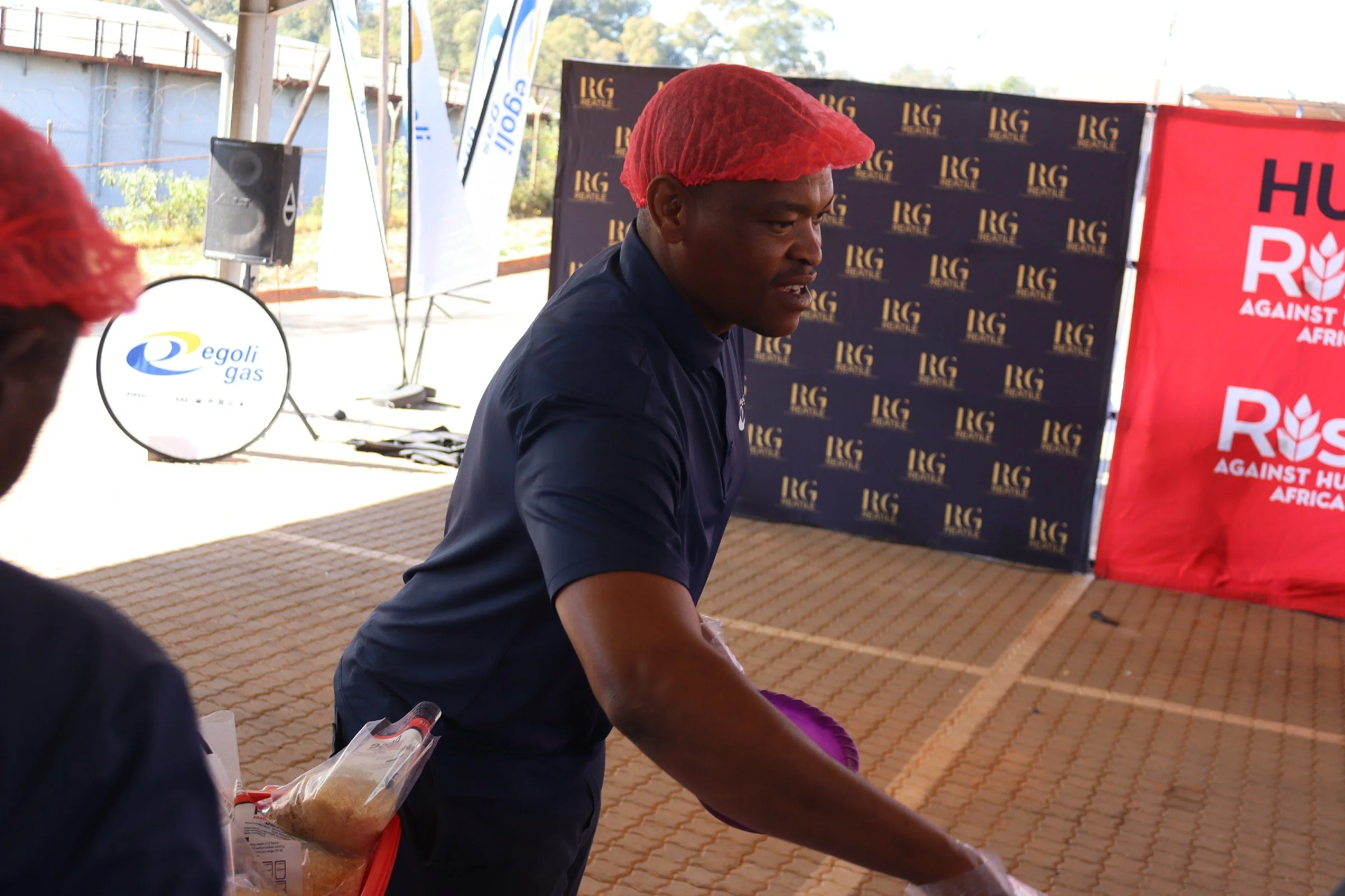 A man wearing a red hairnet and a navy shirt is in an outdoor setting with banners, one red with white text and another black with gold text, and a speaker in the background. He appears to be engaged in an activity related to food or a demonstration.