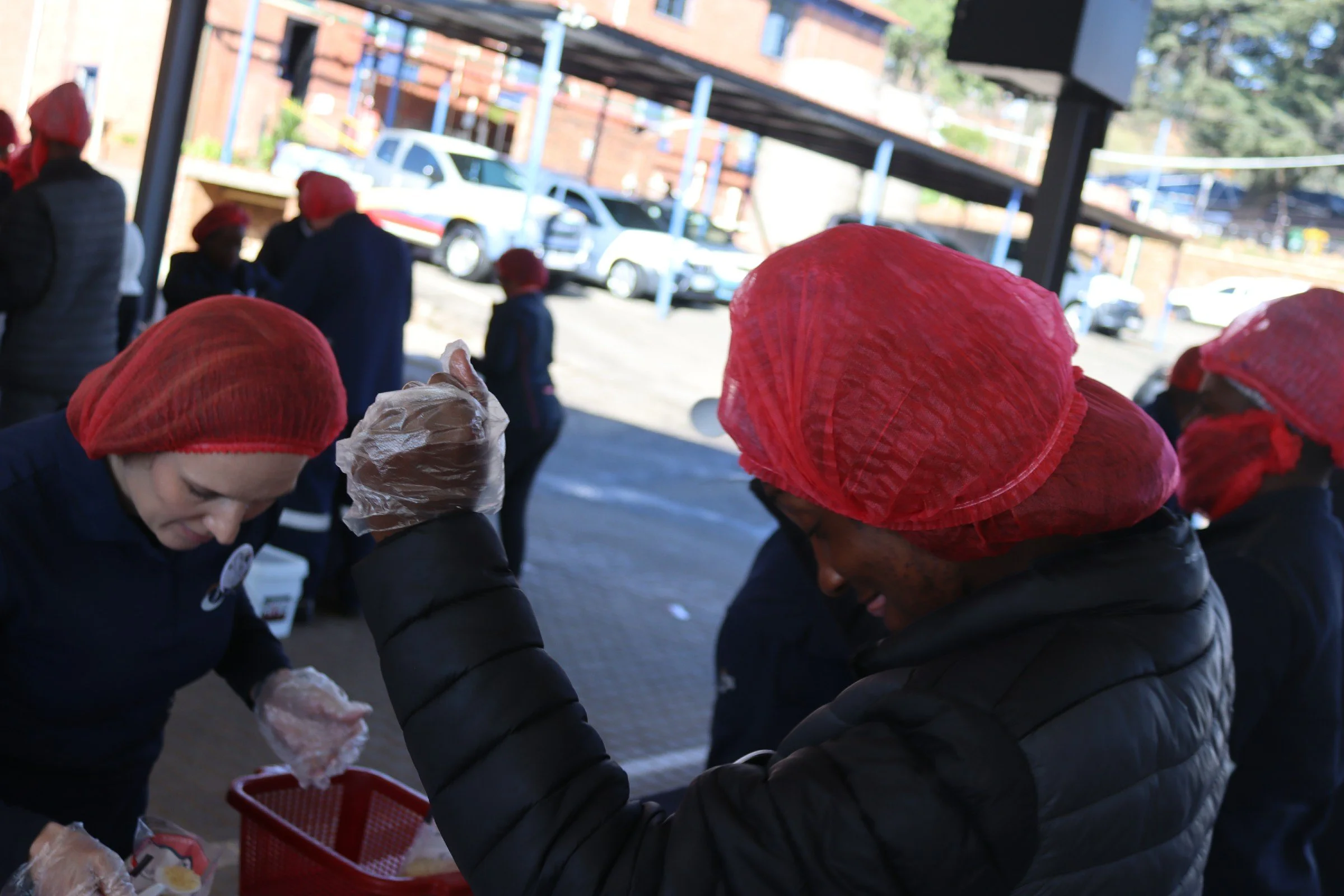 People wearing red hair coverings and gloves working together outdoors, with cars and building in the background.